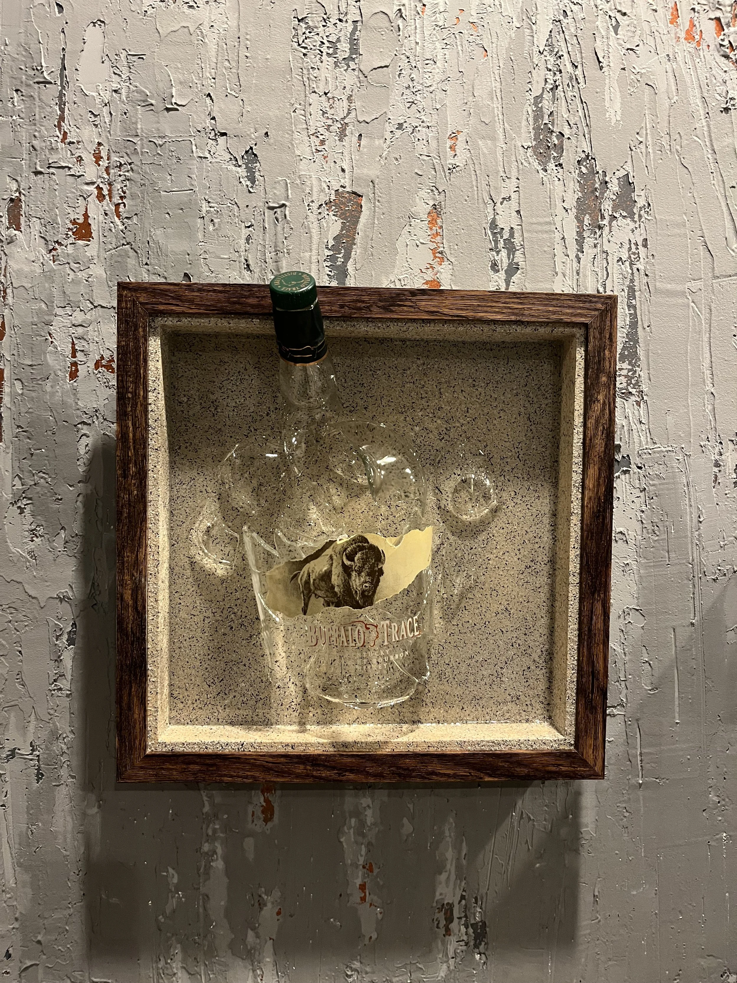 A framed piece of art featuring a glass of alcohol with a dog decal on it, mounted on a textured wall with peeling paint.