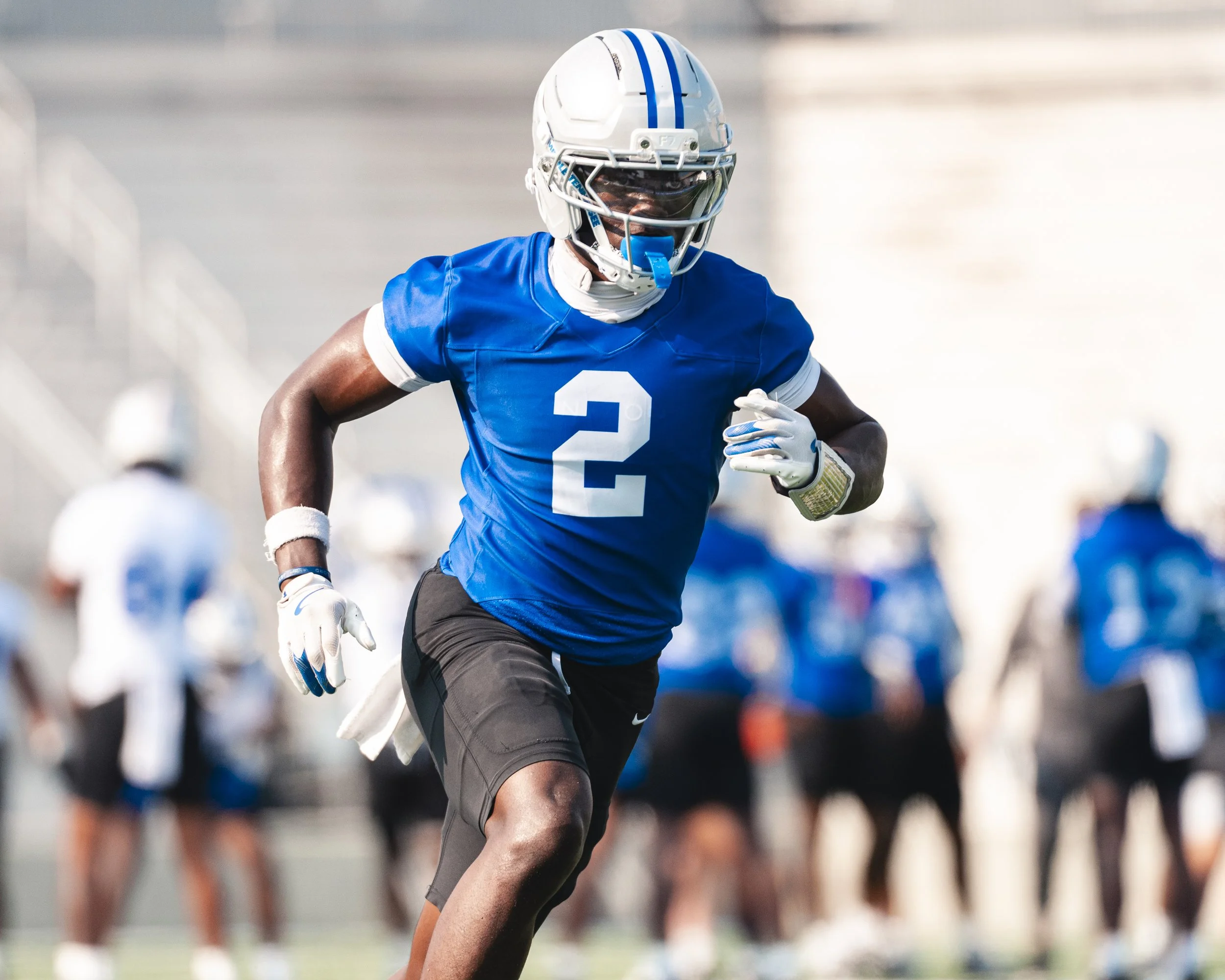 A football player wearing a blue uniform and helmet with the number 2 on it, running on the field during a game with other players blurred in the background.