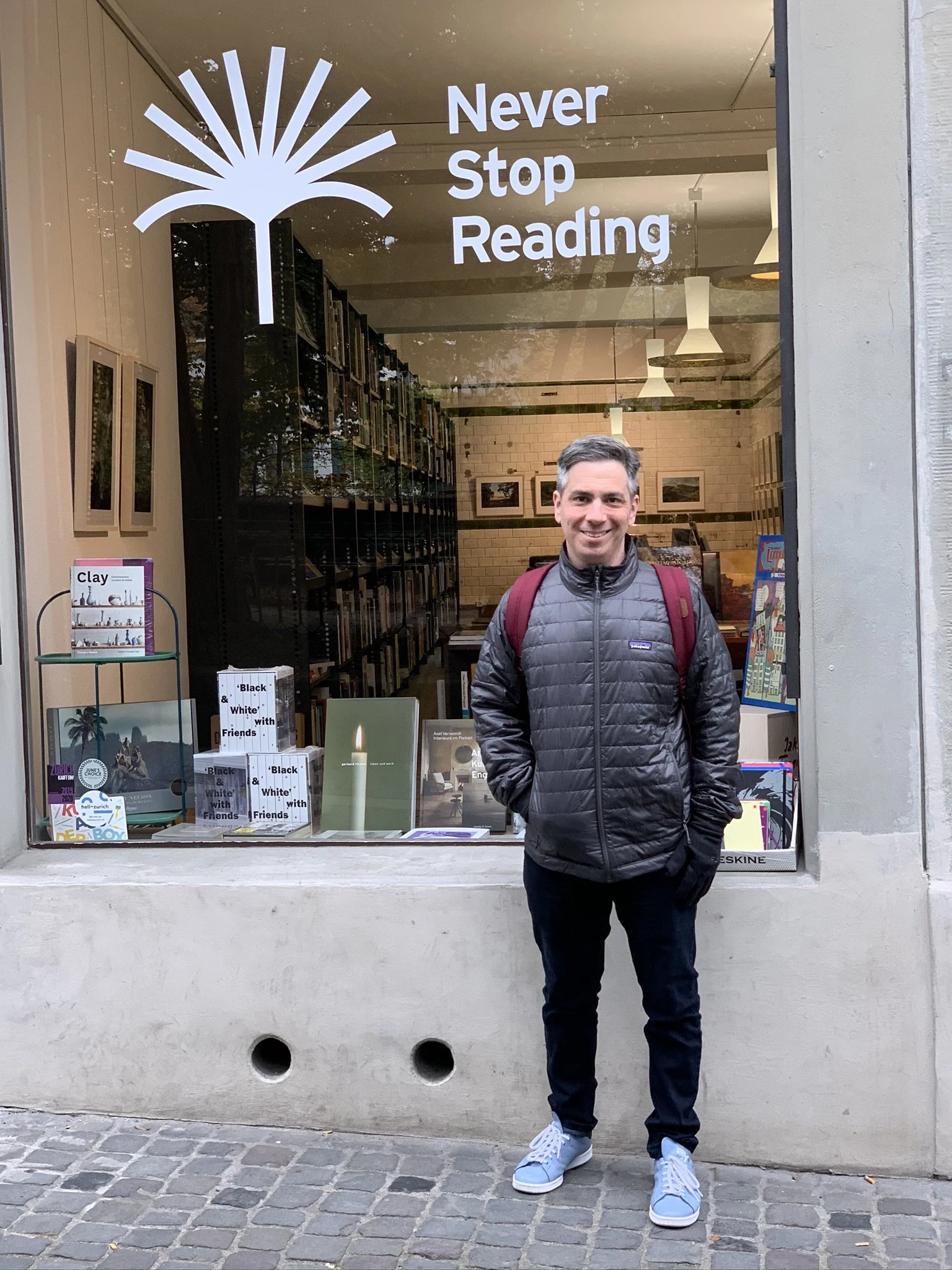 A smiling man with a backpack standing in front of a bookstore window that has the phrase 'Never Stop Reading' on it.