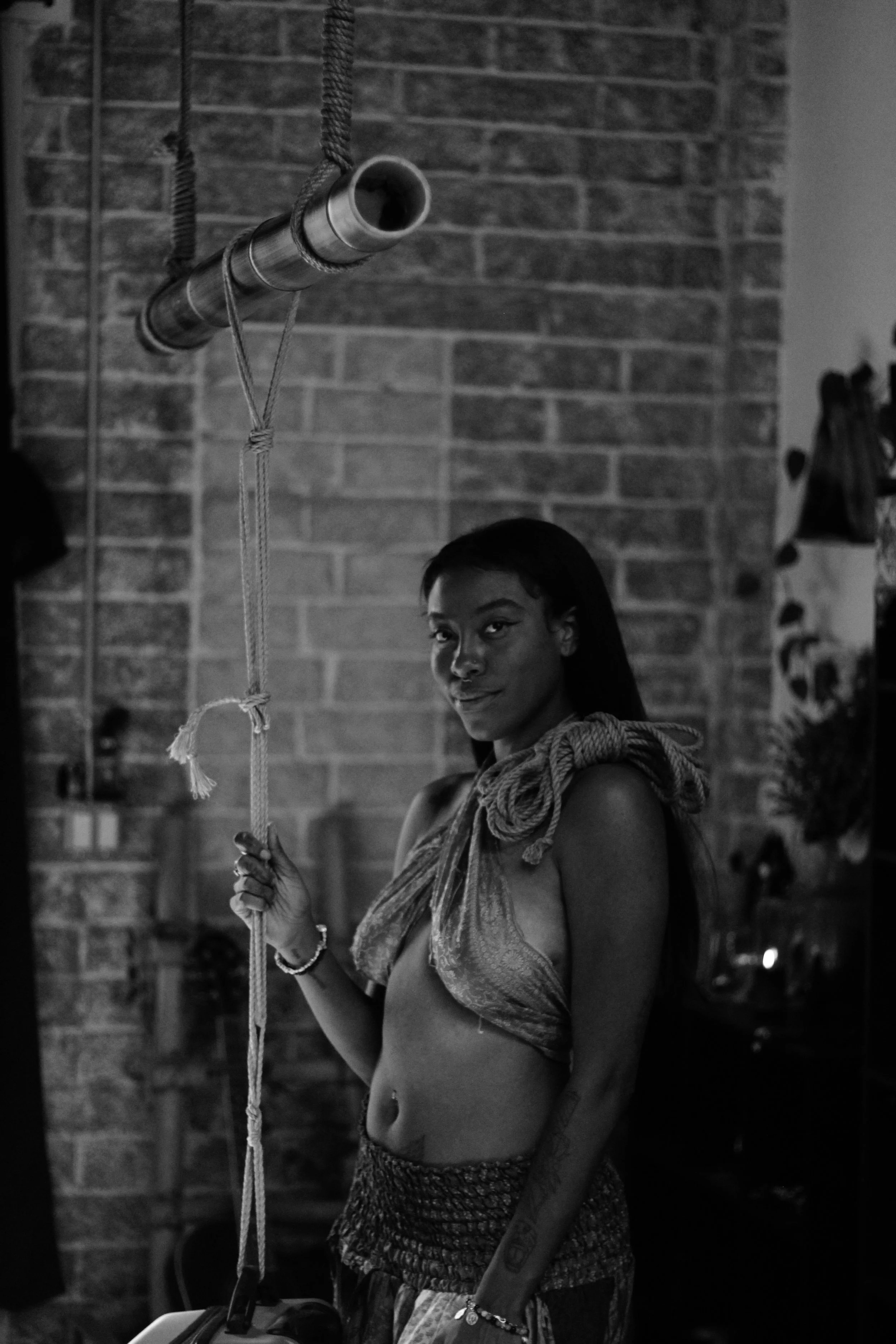 A woman with long dark hair and tattoos on her arm, wearing a patterned top and shorts, holding a rope in her hand, standing indoors with a brick wall in the background.