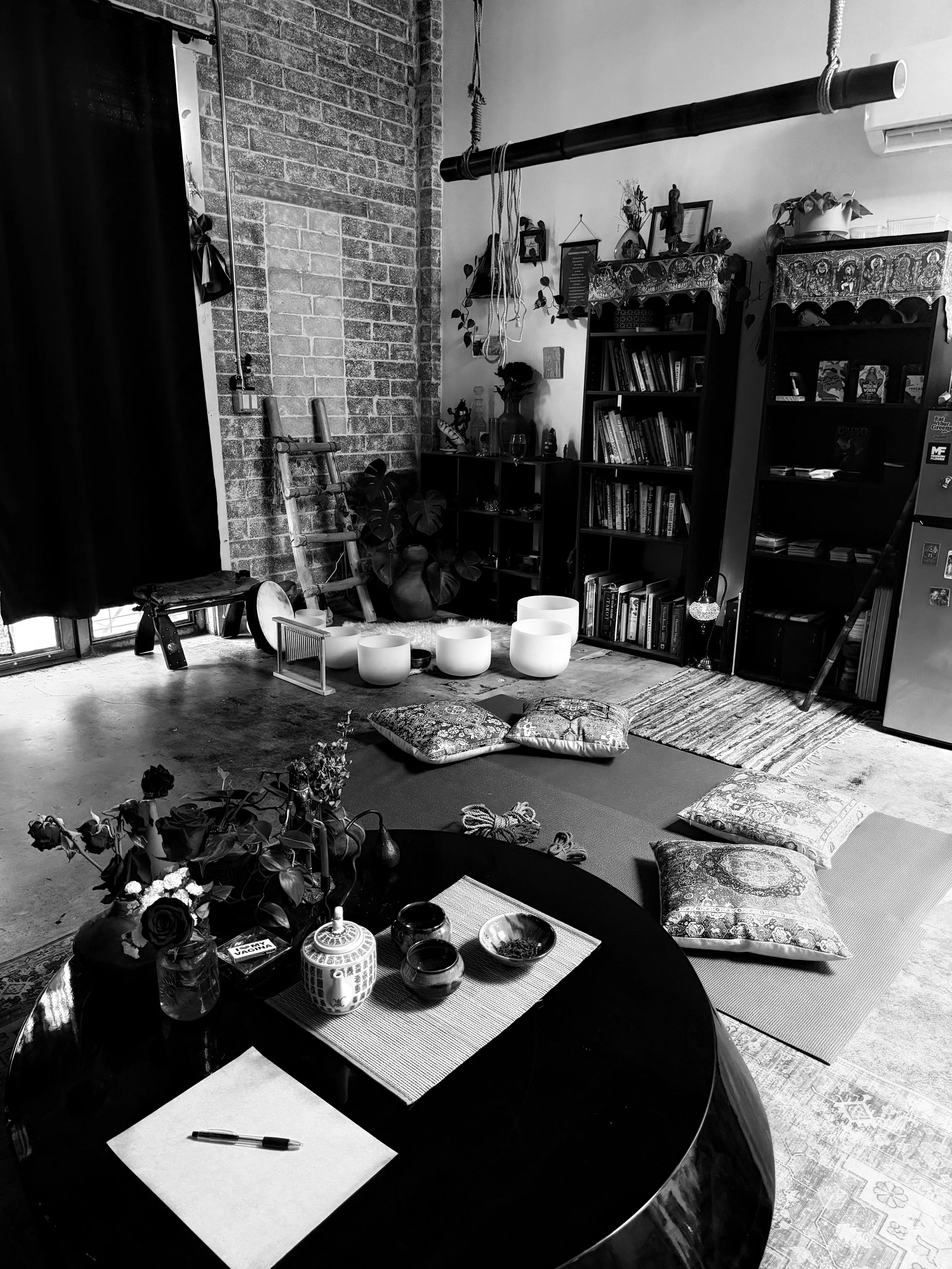 A cozy living room with a black round table set with a notebook, pen, and decorative bowls, surrounded by floor cushions, a bookshelf filled with books, decorative items, and a plant, with a brick wall and hanging ropes in the background.