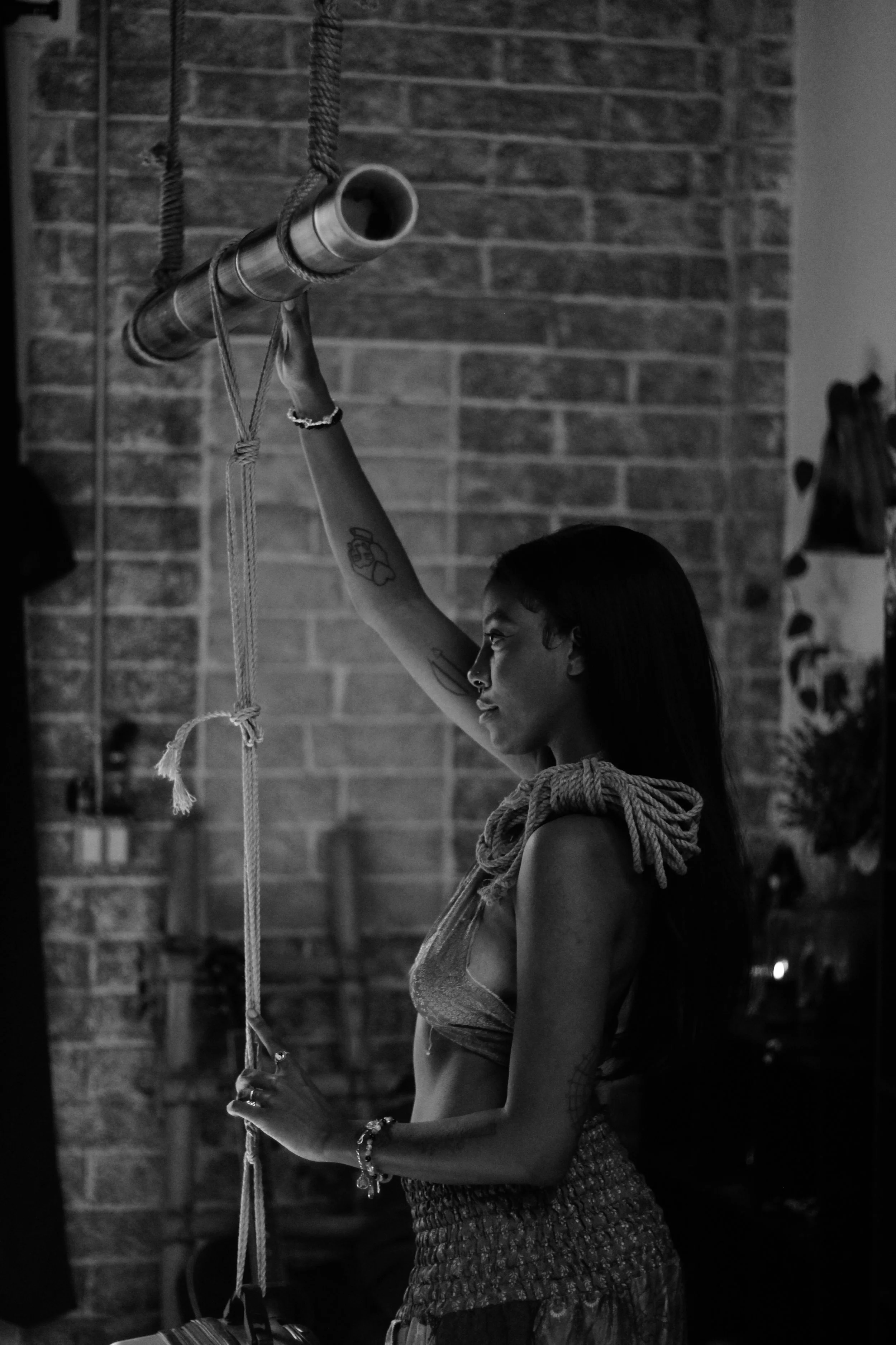 A young woman with long hair, tattoos, and wearing a bracelet, is holding onto a pole with both hands, standing in front of a brick wall.