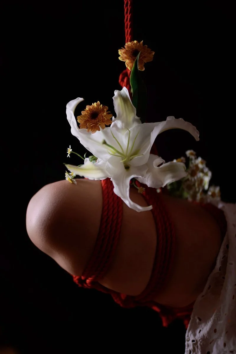 A floral arrangement with white lilies, orange flowers, and small white daisies is tied with red string and suspended in front of a dark background, partially resting on a person's knee.