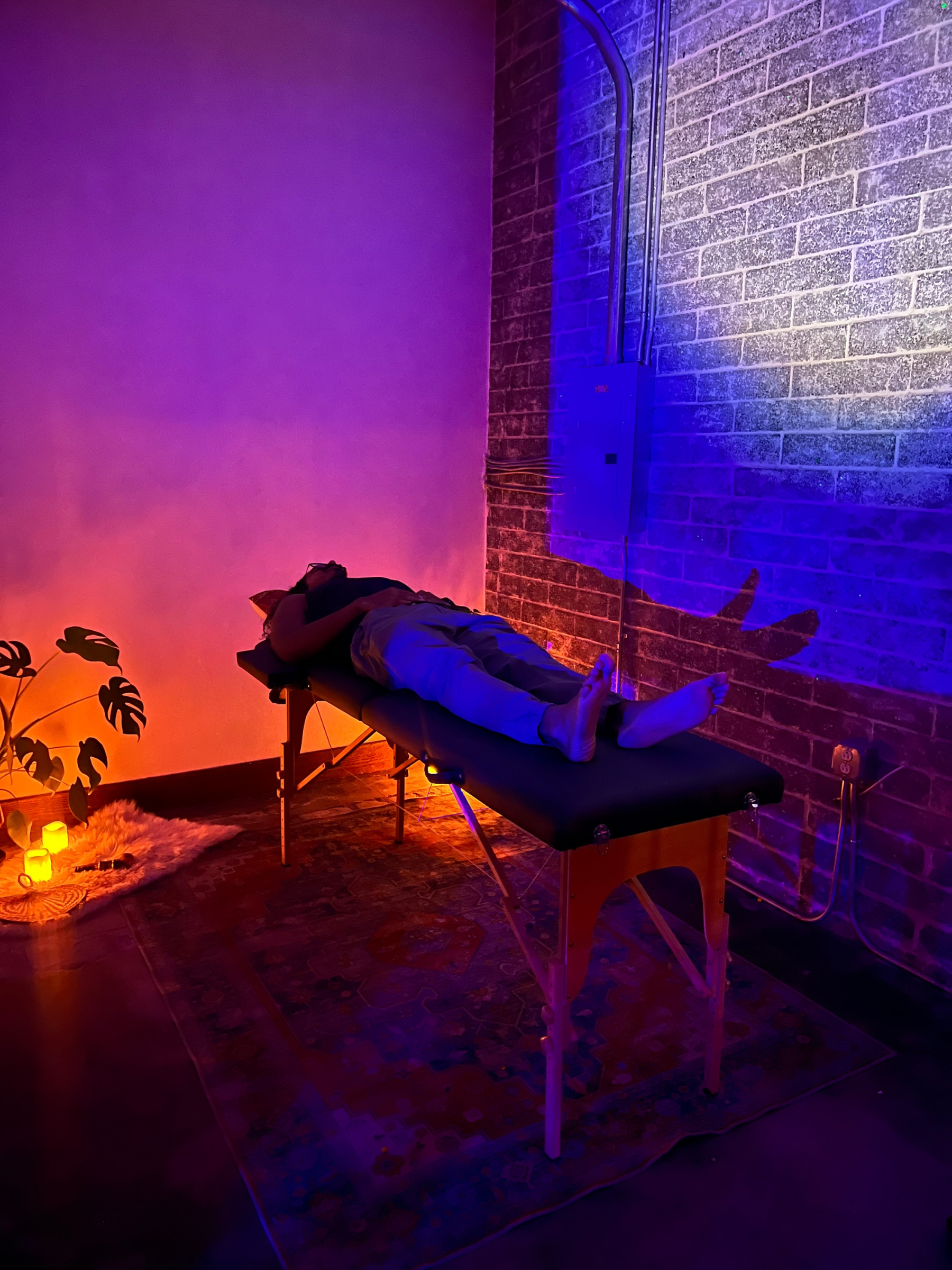 A person lying face down on a massage table in a dimly lit room with purple and orange lighting, brick wall, potted plant, and small lamps on the floor.
