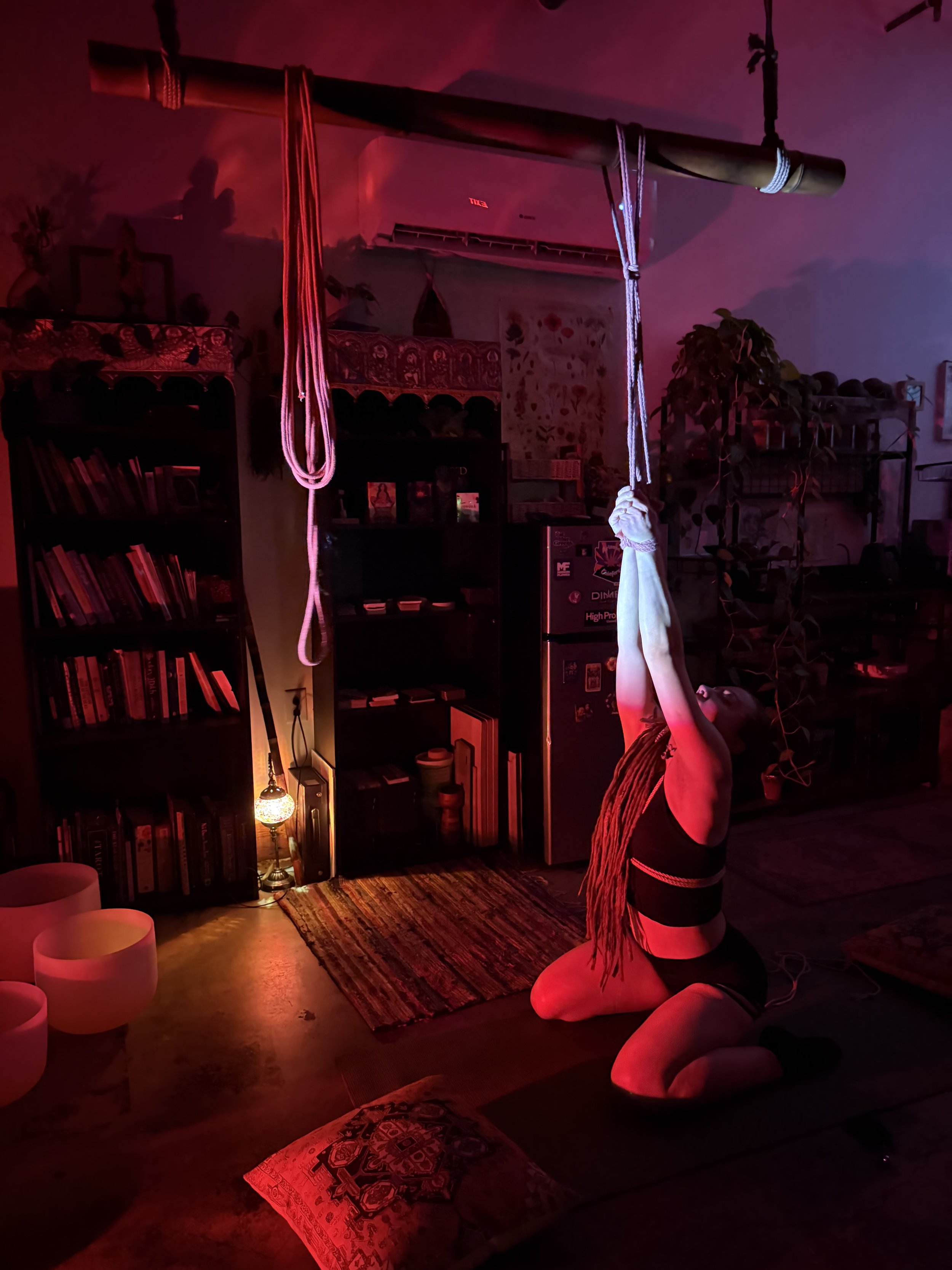 A person with dreadlocks and dark clothing is kneeling on the floor in a dimly lit room illuminated by red and purple lights. They are reaching upward, holding onto a tie rope hanging from a wooden bar attached to the ceiling. The room contains books