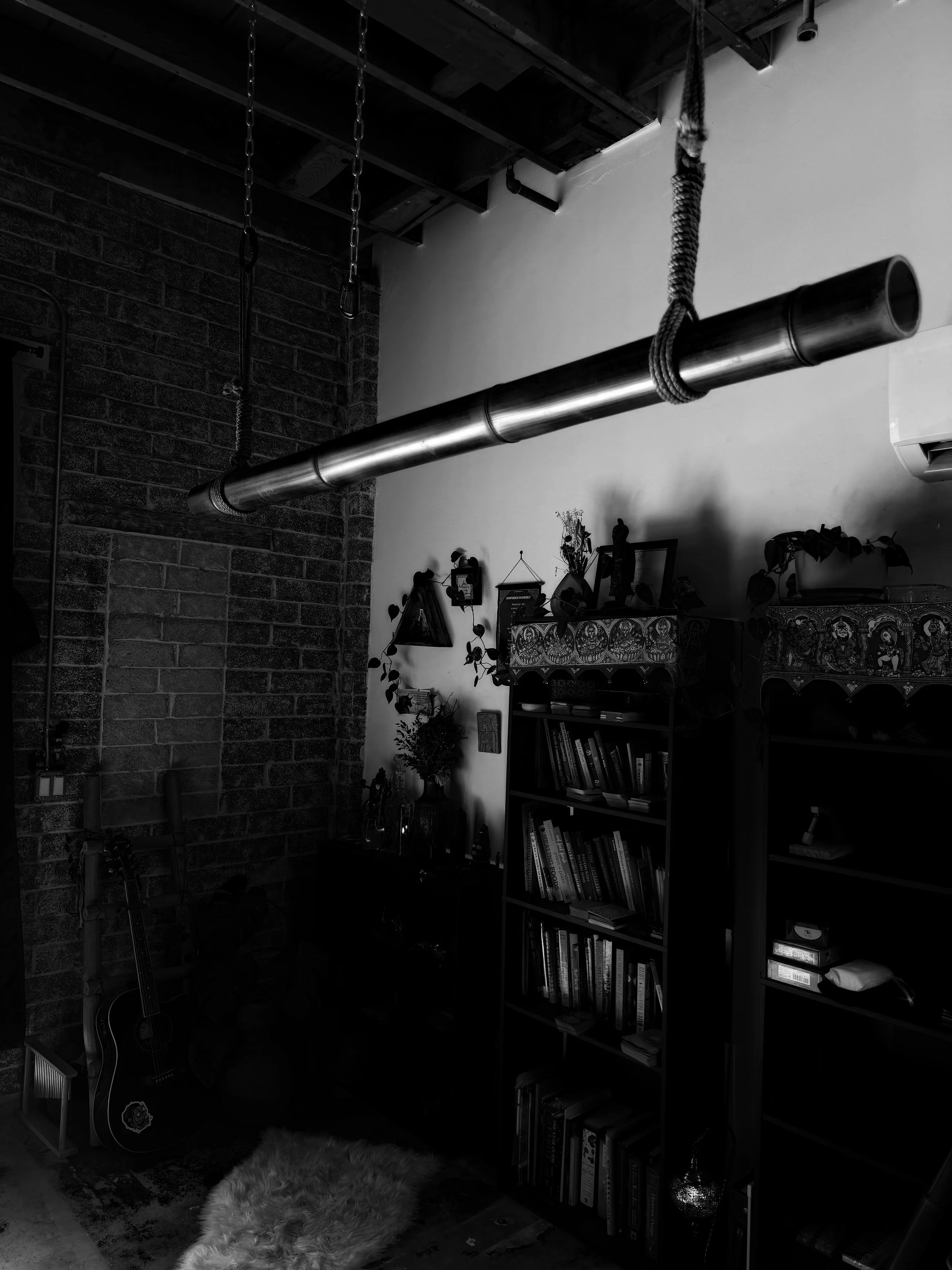 A black-and-white photo of an interior room with exposed brick and wooden ceiling beams. A large metal pipe hangs from the ceiling, with a rope looped around it. The room contains a bookshelf filled with books, a guitar leaning against the wall, and various small decorative items and plants. There is a fluffy rug on the floor.