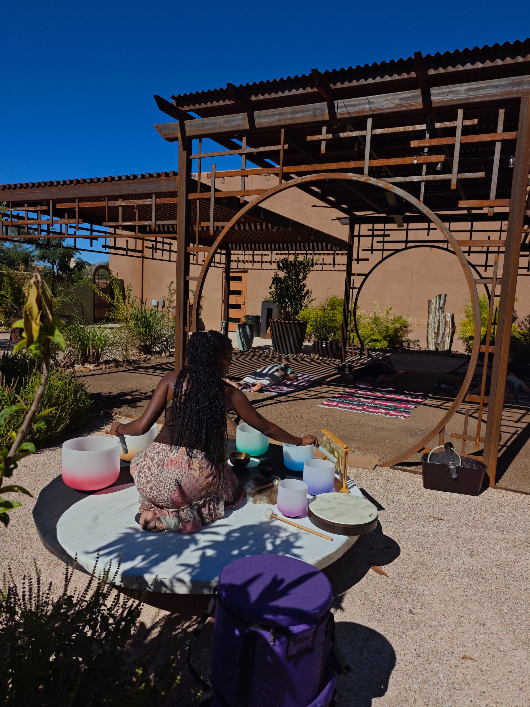 A woman sitting on a large, round, white table playing crystal singing bowls outdoors, with a wooden pergola structure and seating area behind her, and a clear blue sky overhead.