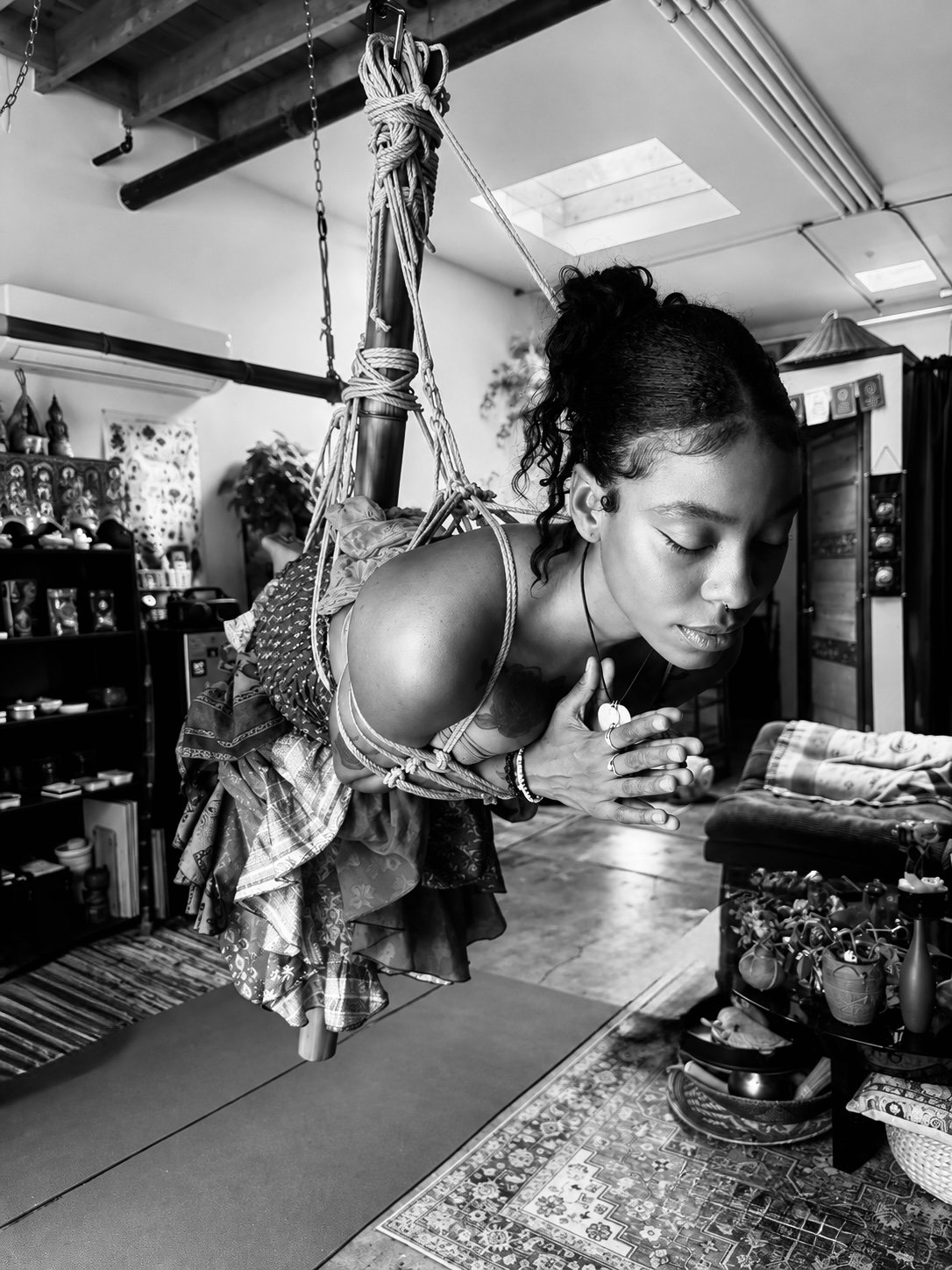 Black and white photo of a woman hanging upside down on a rope in a room decorated with shelves and plants, with her hands folded in prayer.