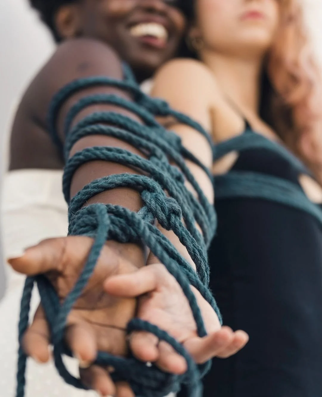 Close-up of two people with their arms wrapped together with numerous blue ropes, smiling and leaning close, highlighting connection or unity.