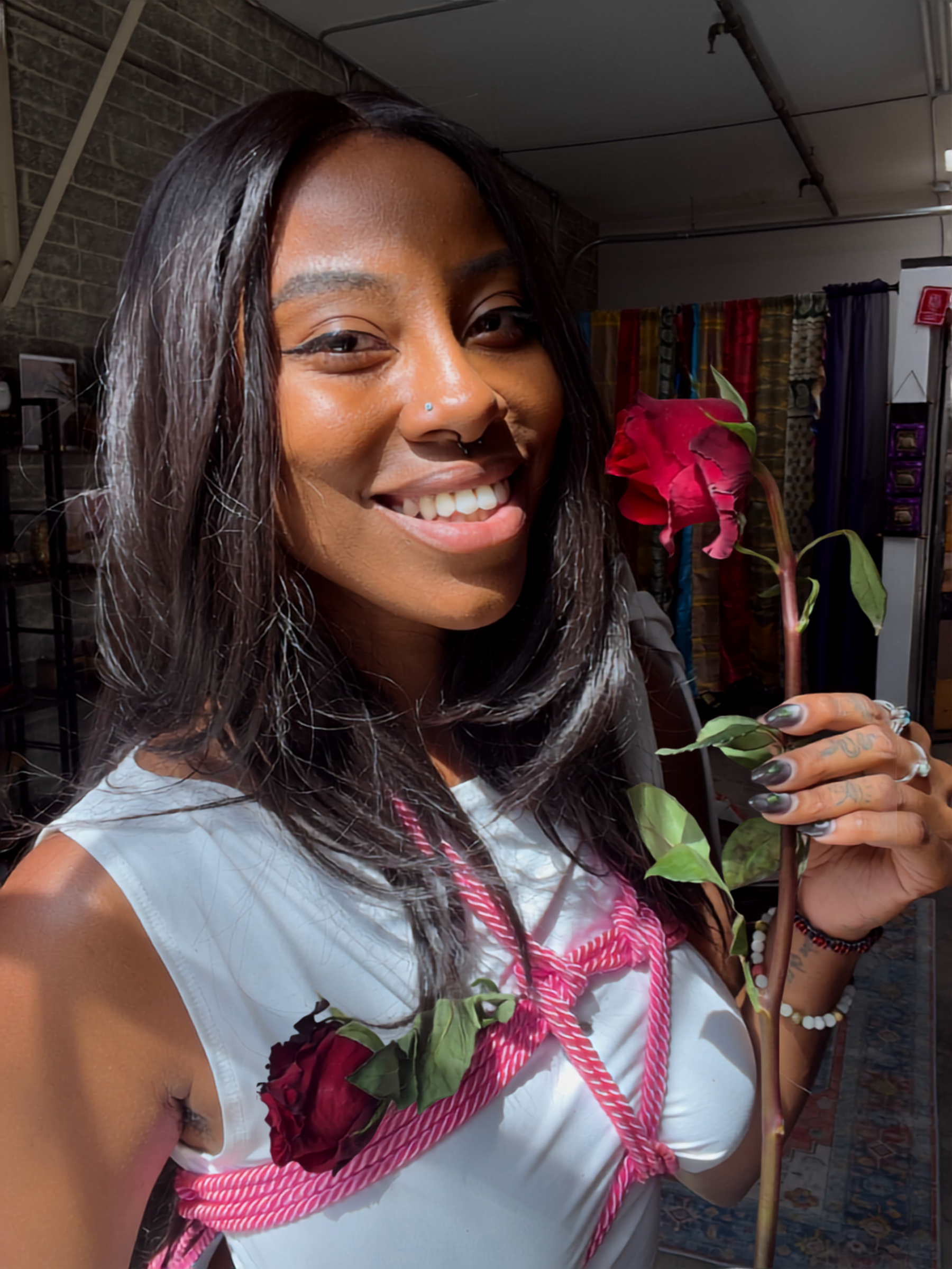 A woman with long dark hair, smiling, holding a red flower, wearing a white sleeveless top, in an indoor space with brick walls and colorful curtains.