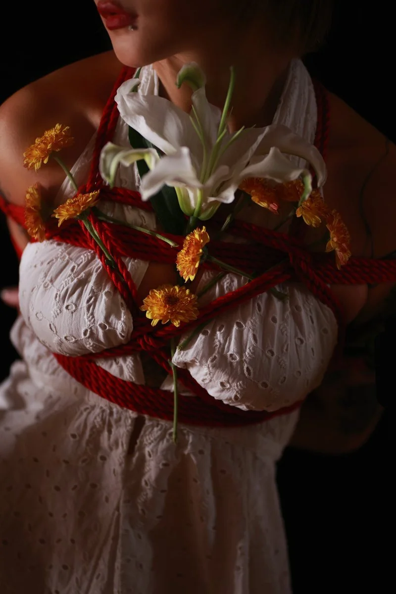 A person in a white embroidered dress with flowers and red rope tied around their chest, holding a bouquet of white lilies and orange chrysanthemums.