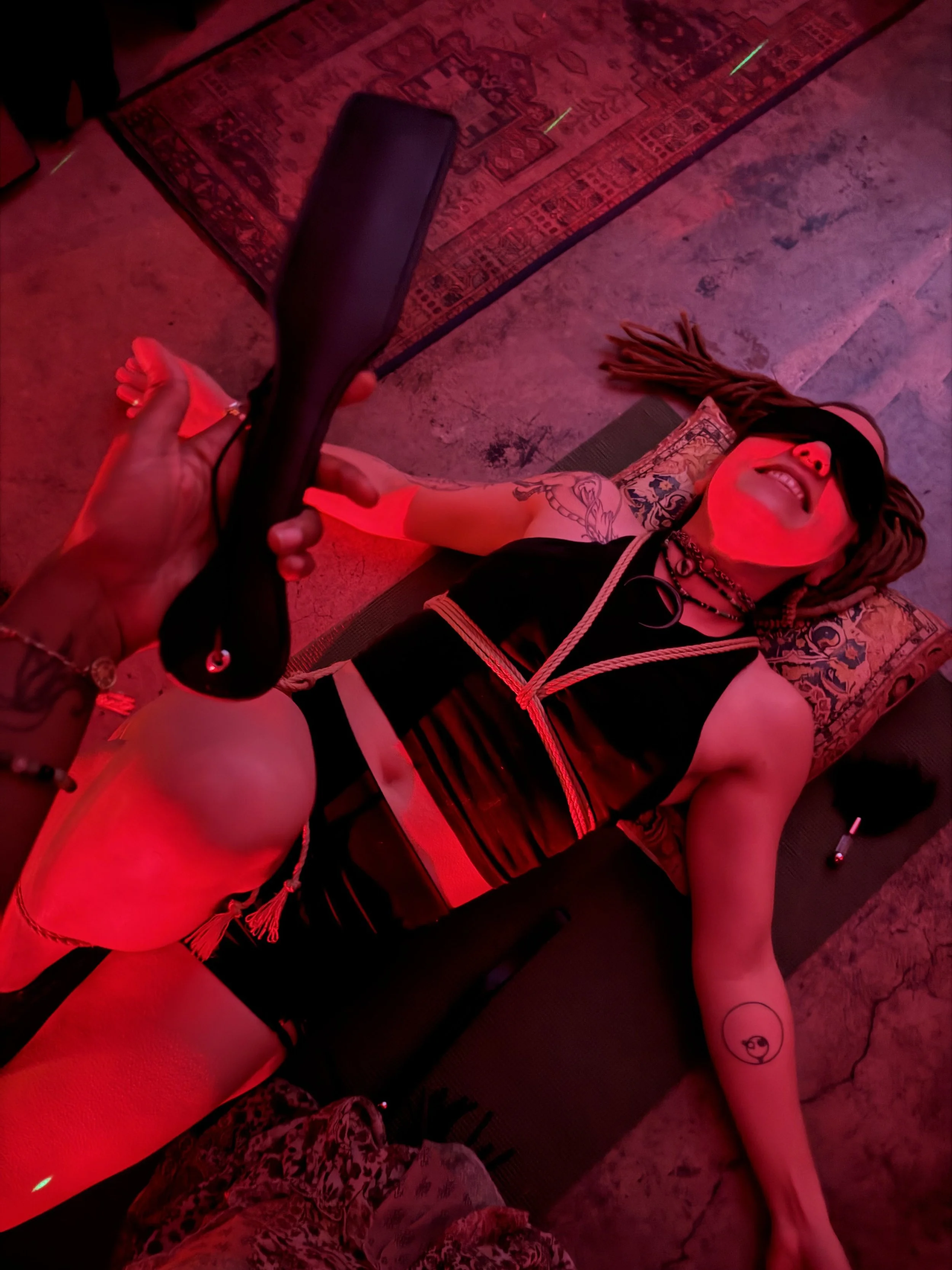 Person lying on the floor, holding a leather whip, with tattoos on their arm, smiling with eyes closed, in a room with red lighting and a patterned rug.