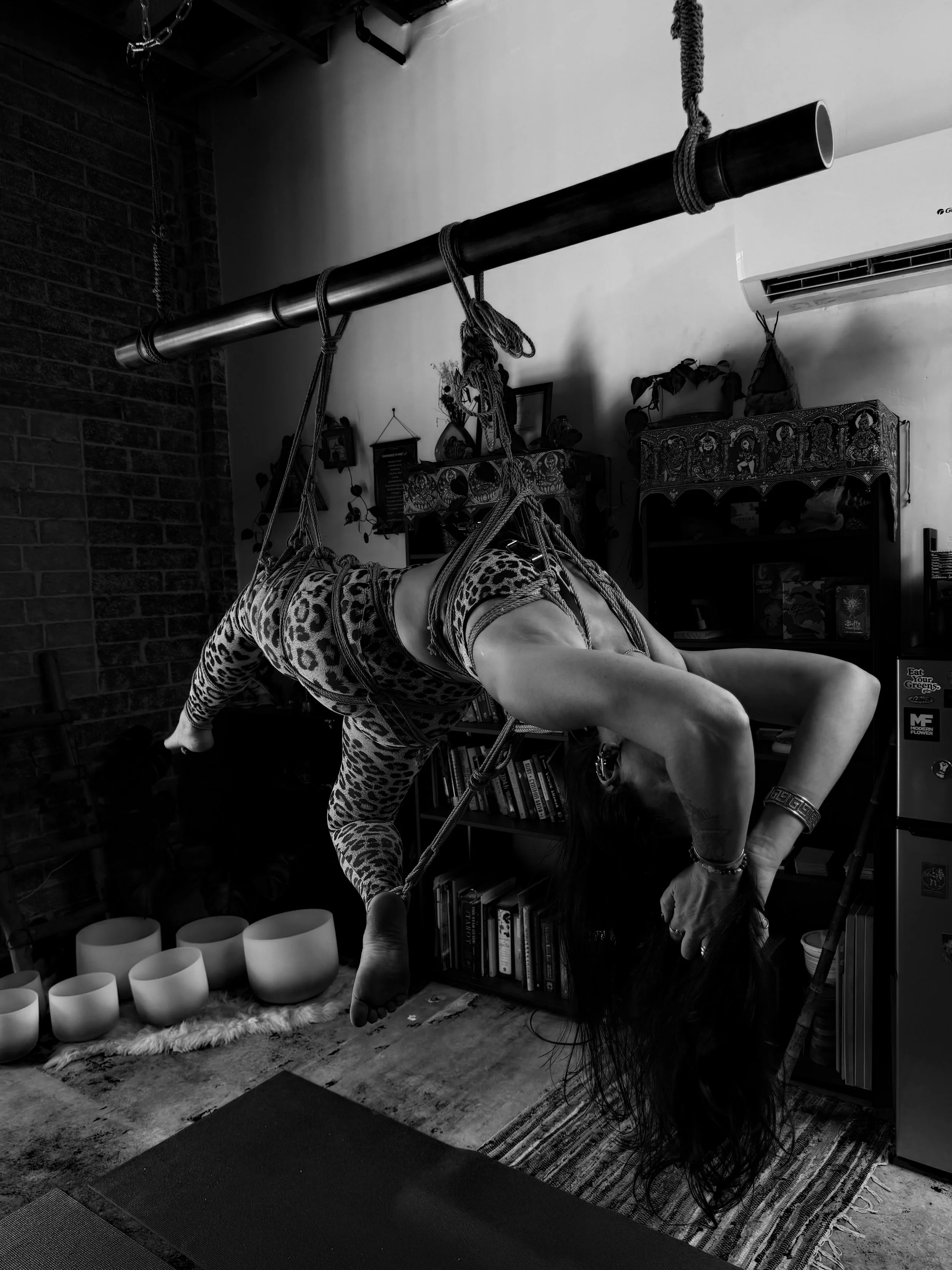 A woman practicing aerial yoga or suspension yoga indoors, hanging upside down from a ceiling-mounted metal bar using ropes or slings, with her head and hair hanging downward and wearing leopard print leggings.