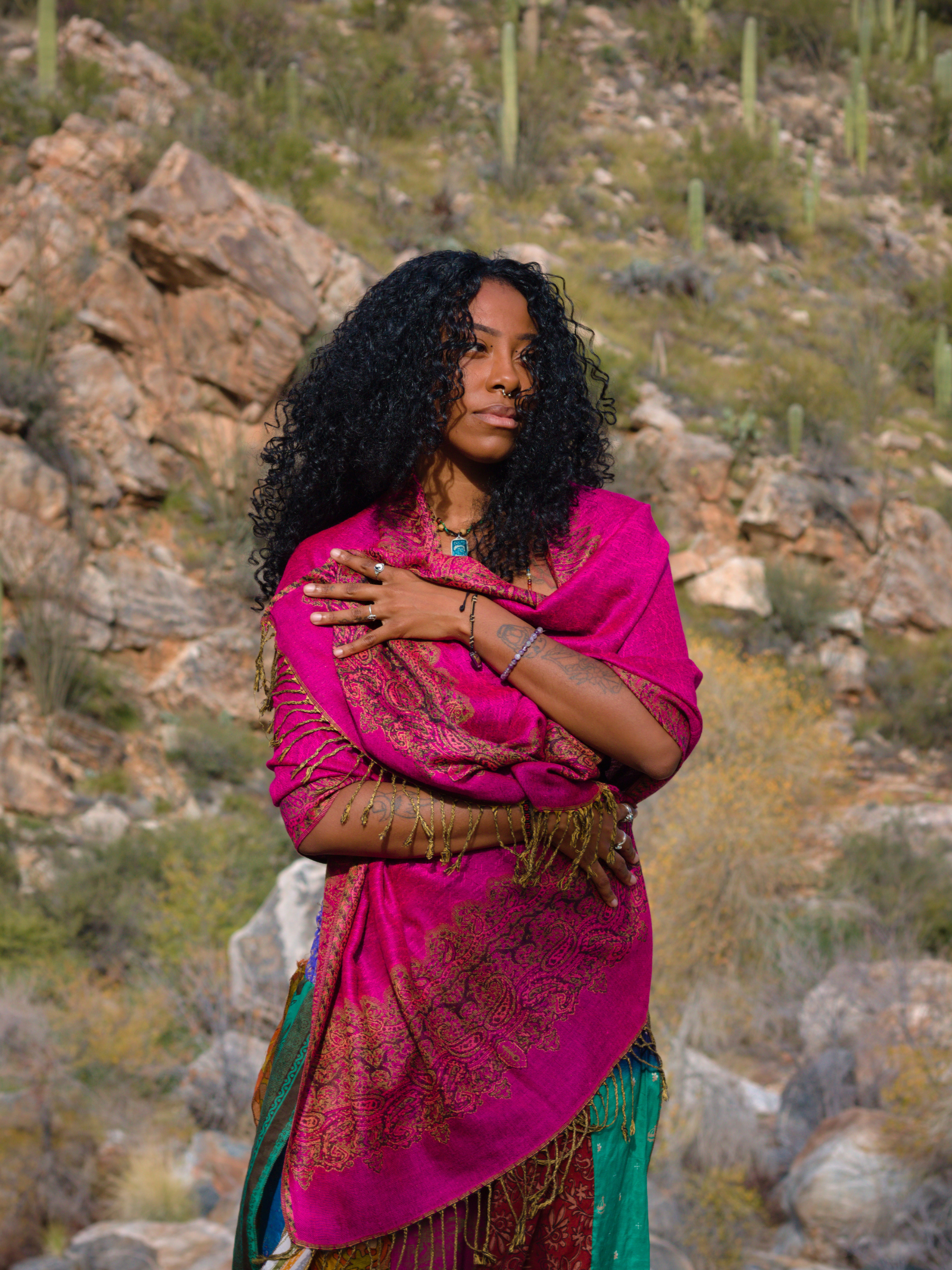 A woman with curly black hair and medium skin tone stands outdoors against a desert landscape with rocks and cactus. She is wearing a vibrant pink shawl with gold embroidery over a colorful dress and has multiple bracelets and rings on her arms and fingers.