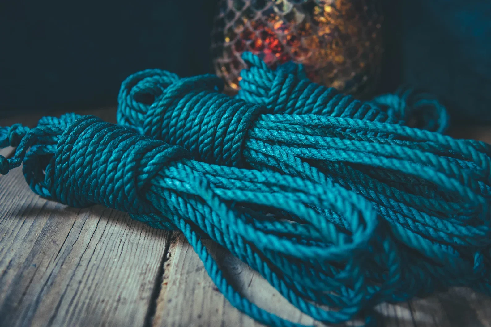 A bundle of teal rope coiled and lying on a wooden surface, with a blurred, multicolored light source in the background.