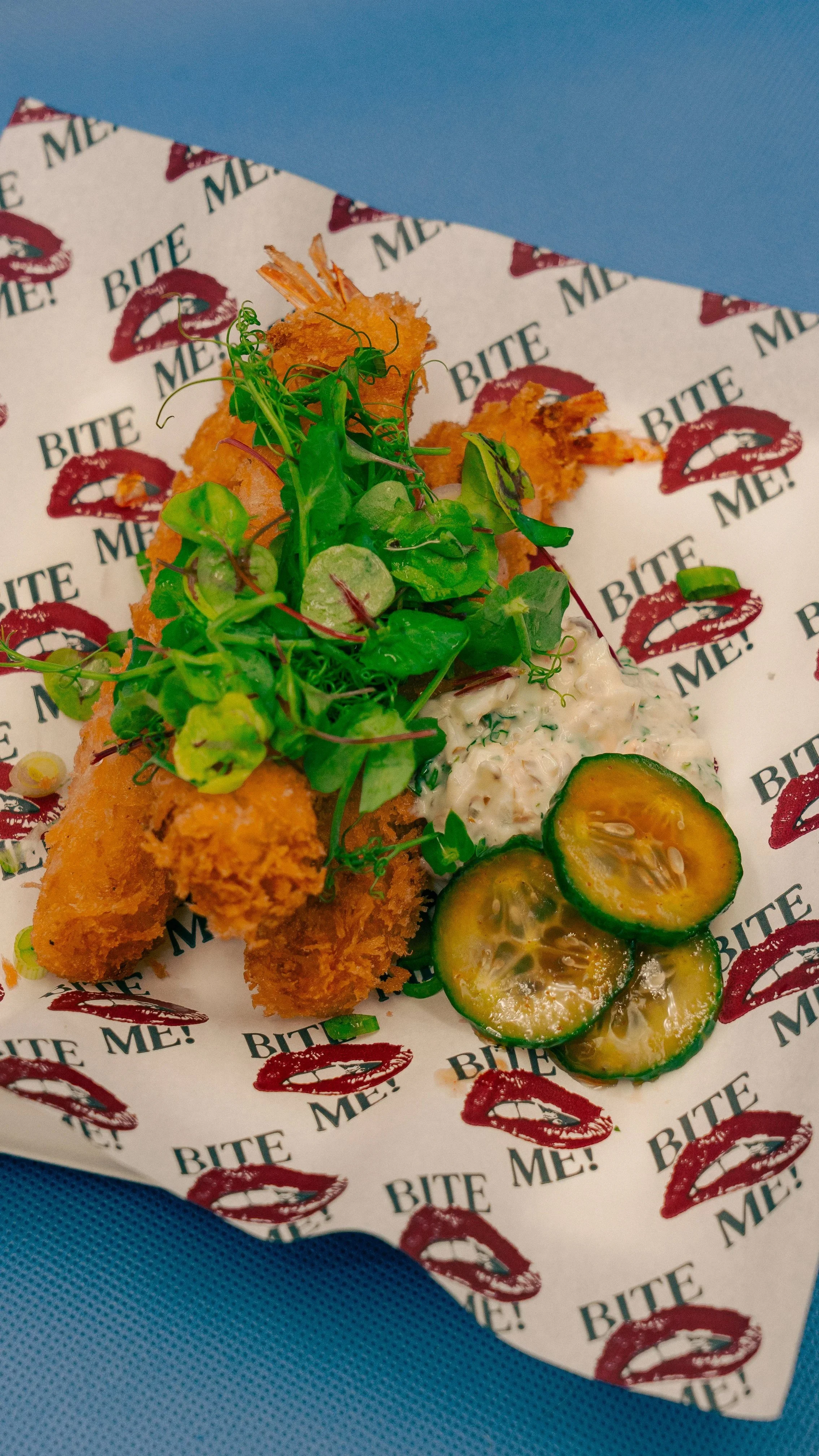 Gefrituurde vis met frisse salade, komkommer en tomaat op bedje van pitabrood, geserveerd op papier met 'BITE ME!' prints.