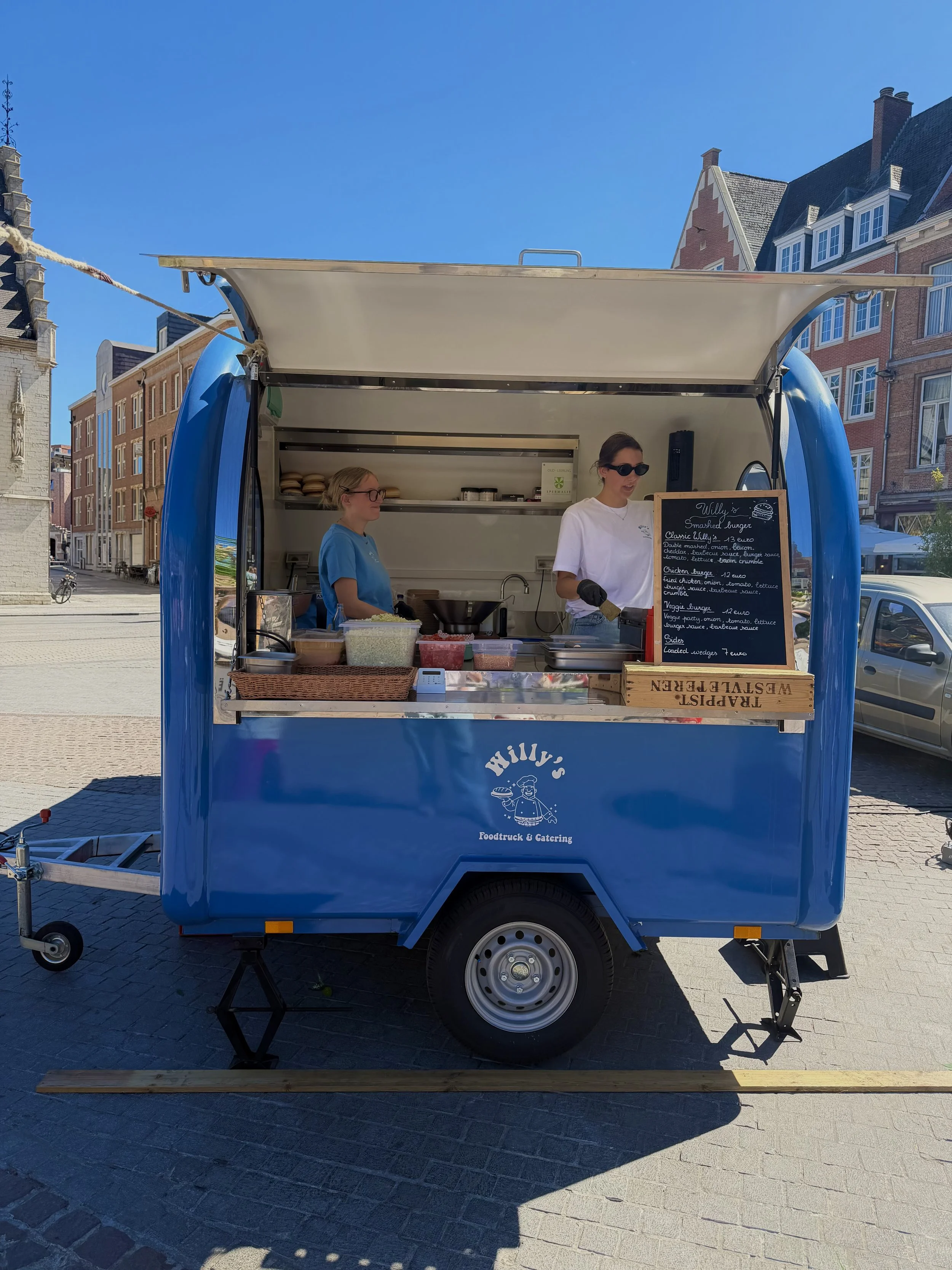 Een blauwe foodtruck met een open keuken en een menu bord, op een straat in een historisch stadscentrum.
