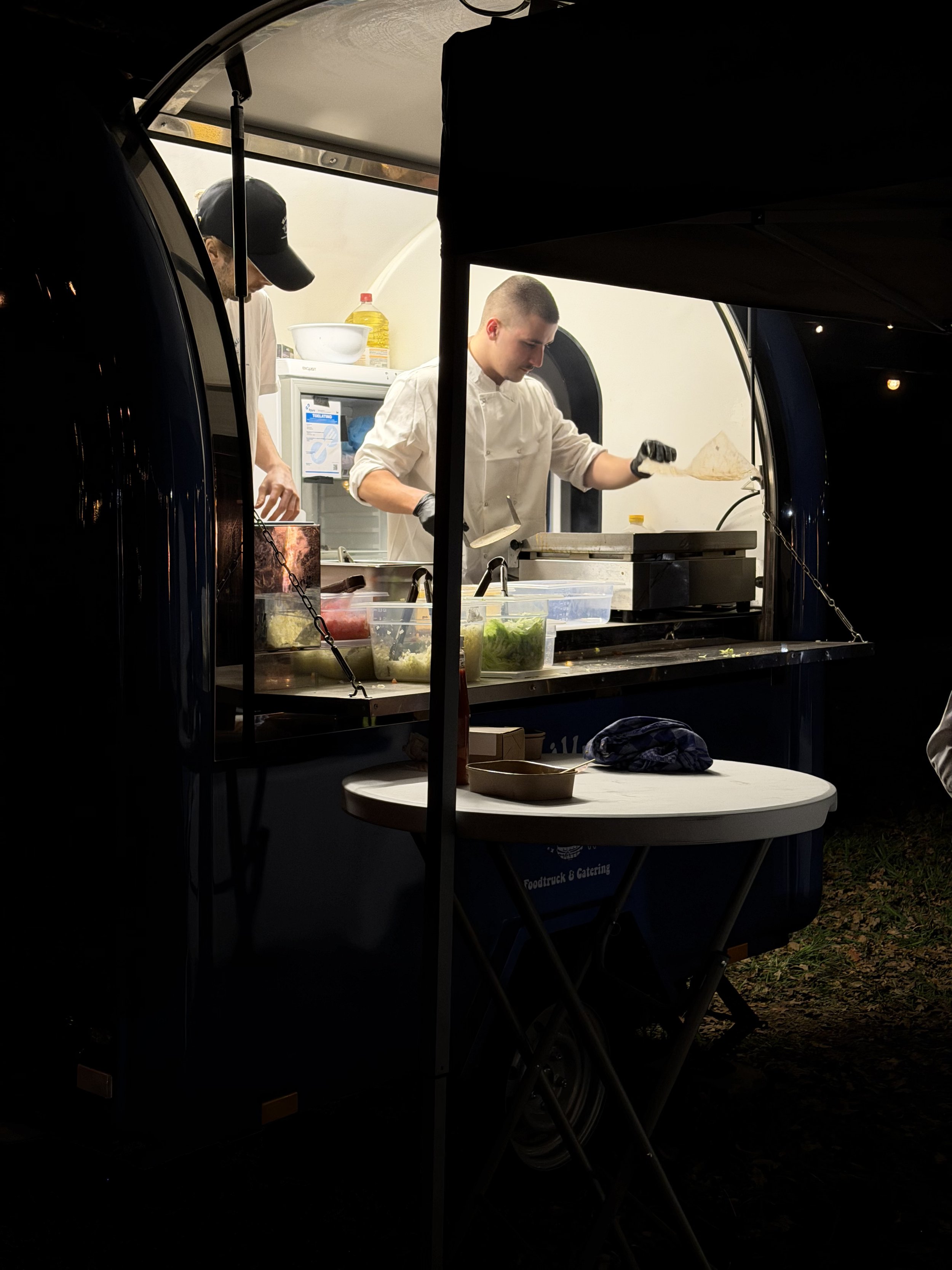 Twee mannen bereiden voedsel in een foodtruck, één man draagt een wit kokssmeker en zwarte handschoenen, de andere man draagt een zwart pet en een lichtkleurig shirt. De voedseltruck is donker en er is een tafel met ingrediënten vastgelegd in de voorgrond.