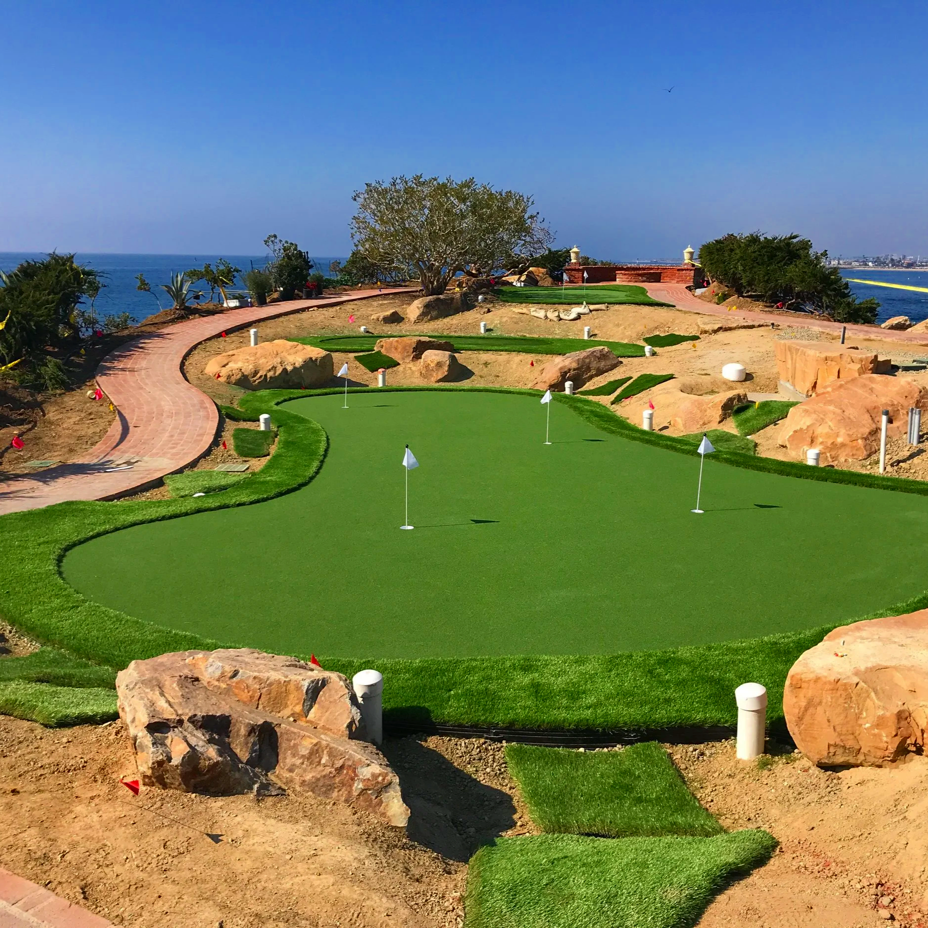 Miniature golf course with green putting greens, flags, and large rocks, situated near the ocean with a city skyline in the distance under clear blue skies.
