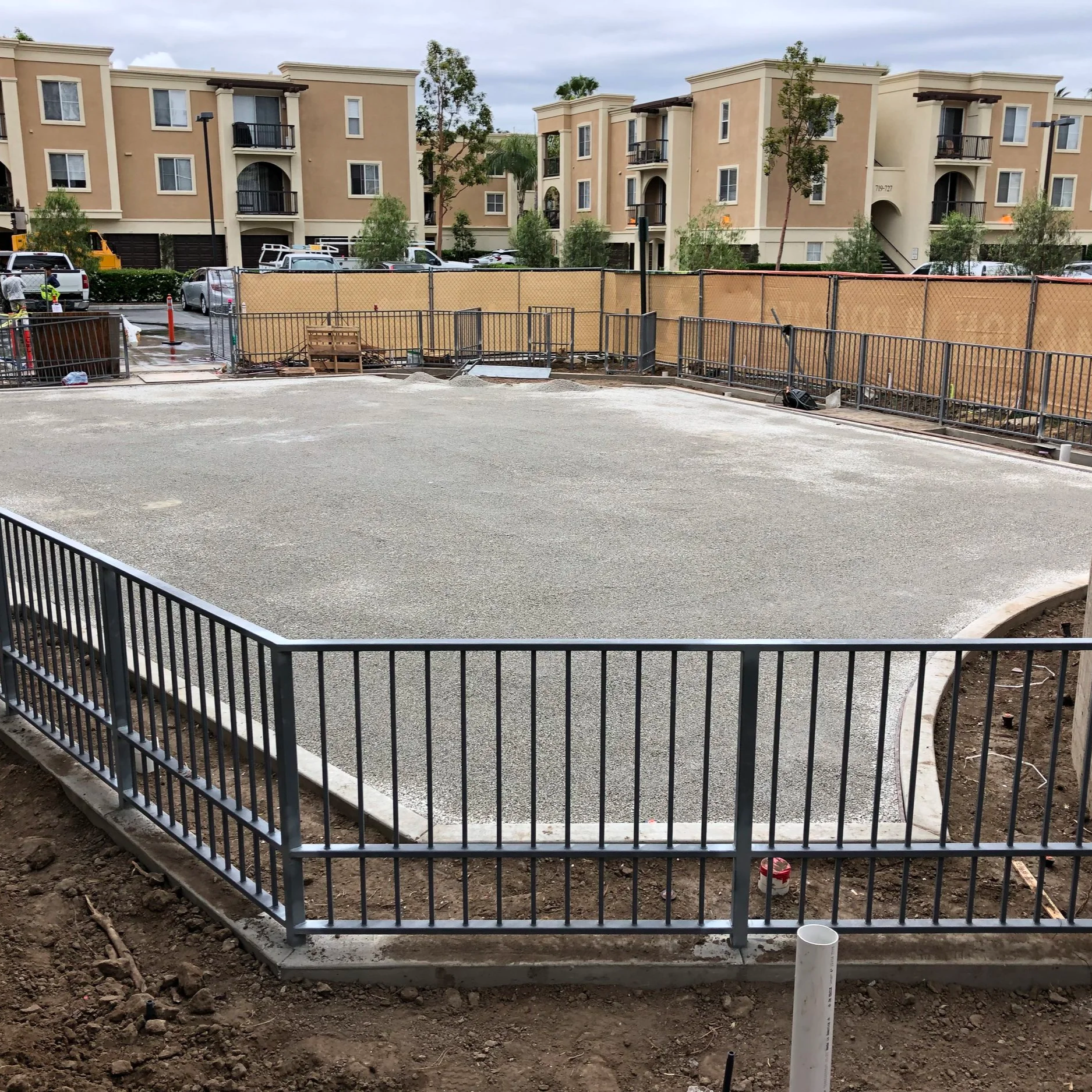 Construction site with a fenced-off area and an apartment complex in the background, with parked cars and trees.