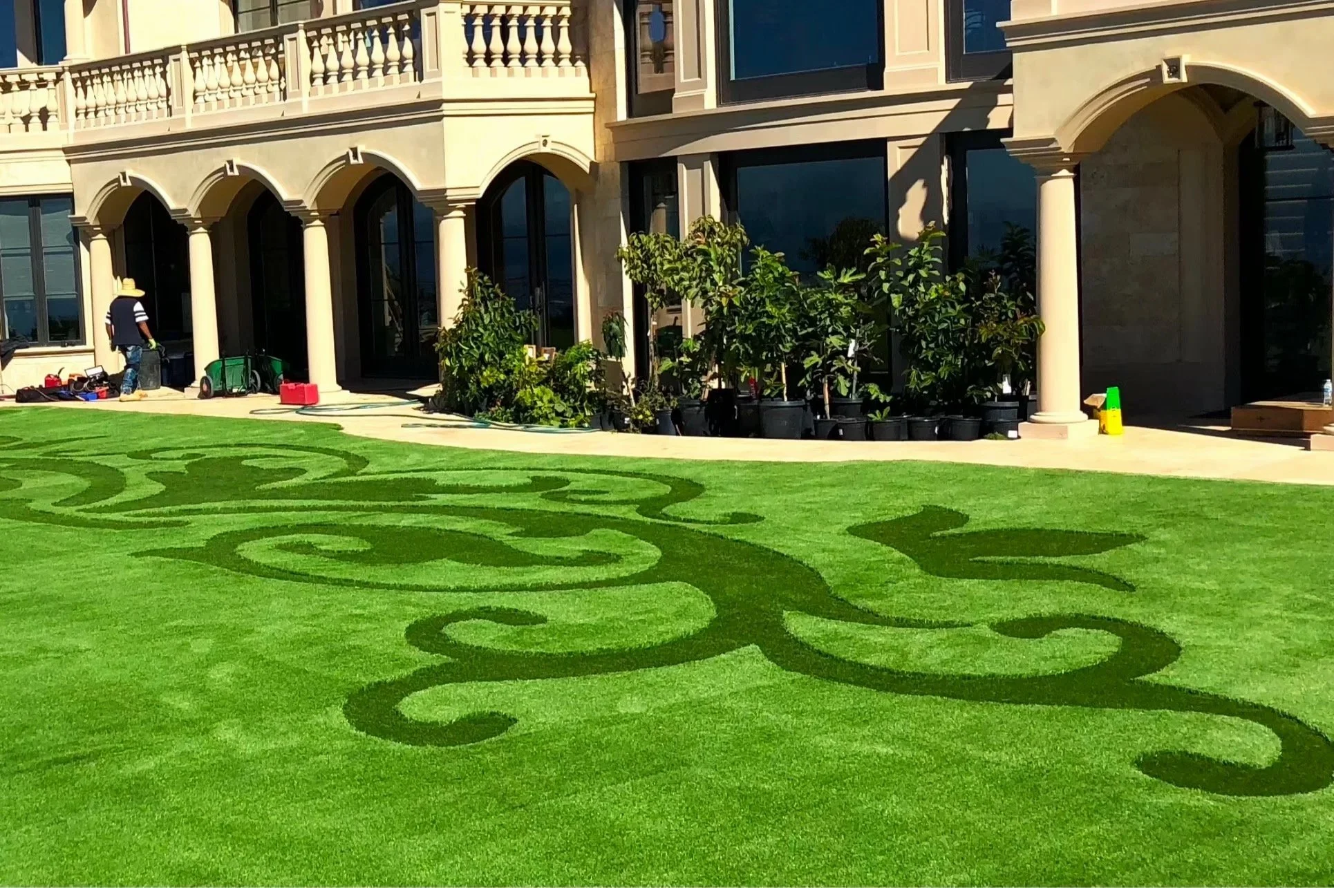 The image shows a large, intricately patterned green lawn in front of a grand house with beige stone exterior, arched doorways, columns, and a balcony. There are potted plants along the house's wall and a person working near gardening tools.