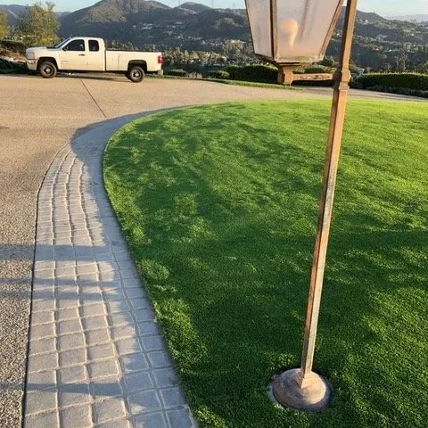 A curved paved pathway next to a well-maintained grassy area with a white truck parked nearby, a traffic sign, and distant mountains in the background.
