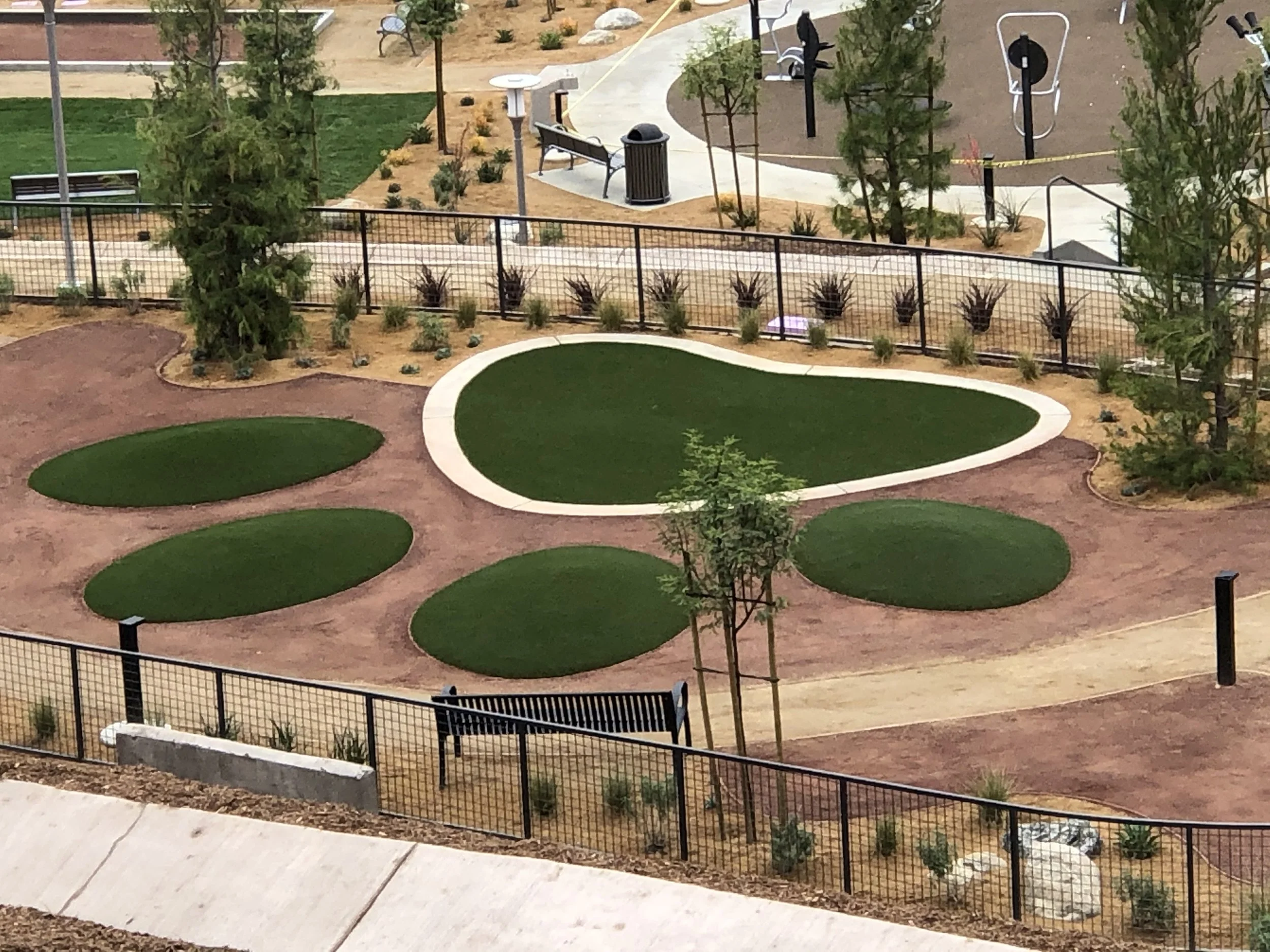 Dog park with an green artificial grass paw print design in decomposed granite with trees, a footpath, bench. Surrounded by plants, trees and a black metal fence