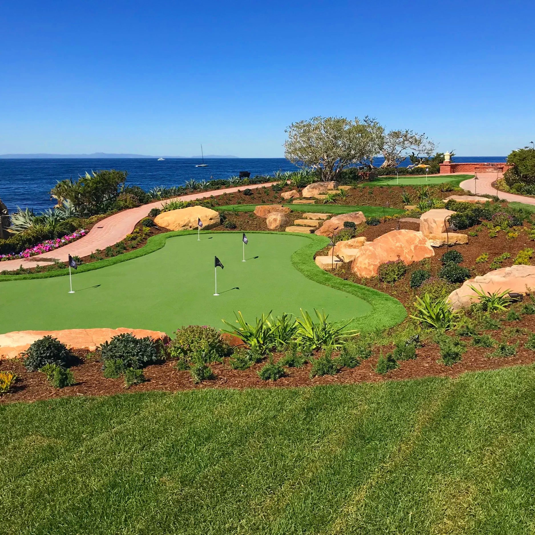 A scenic coastal garden with a putting green, surrounded by rocks, plants, and flowers, overlooking the ocean with sailboats and a distant landmass.