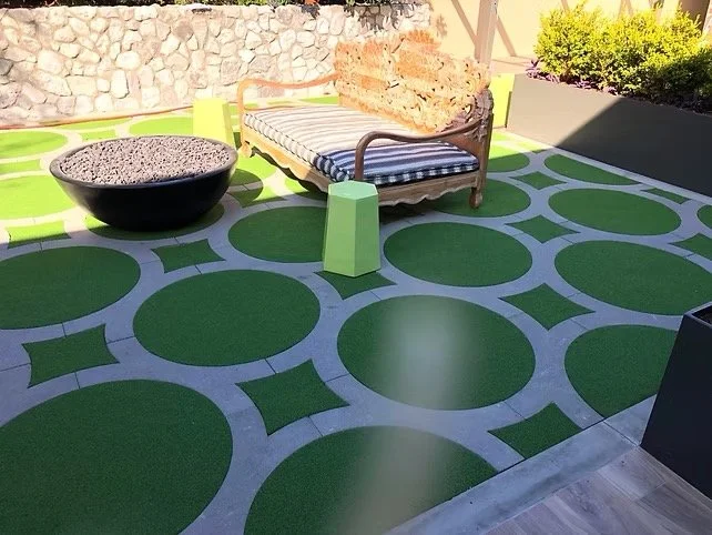Outdoor patio with a green and gray patterned floor, a black firepit filled with gray stones, a wooden bench with striped cushions, and small green side tables. There are bushes and a stone wall in the background.