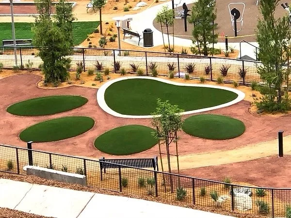 An empty playground with artificial grass in various shapes surrounded by a black metal fence, with young trees and shrubs planted around the area.