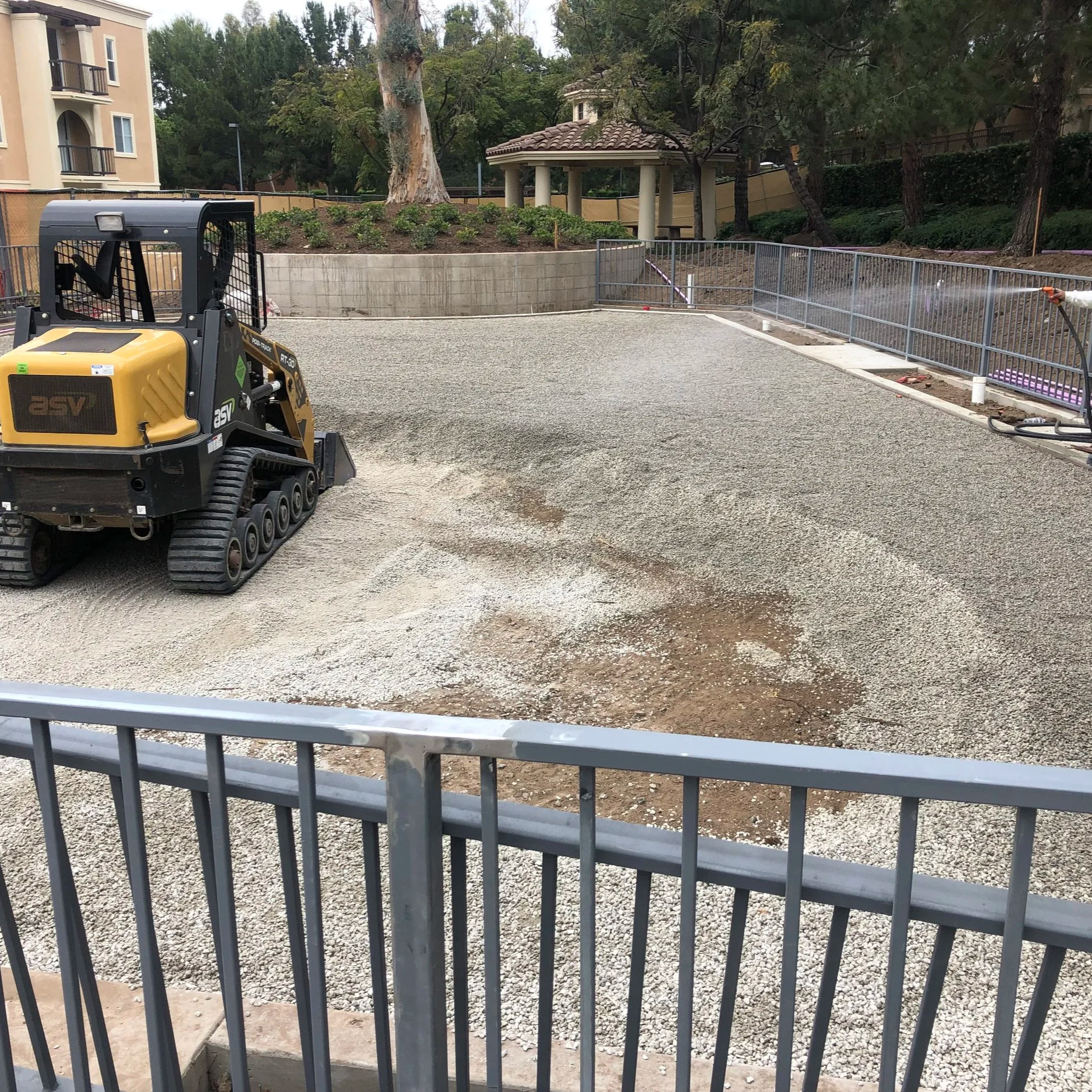Construction site with gravel ground, metal fencing, and a small yellow skid-steer loader near the foreground. There is a landscaped area with trees and shrubs, a gazebo with columns, and a residential building with balconies in the background.