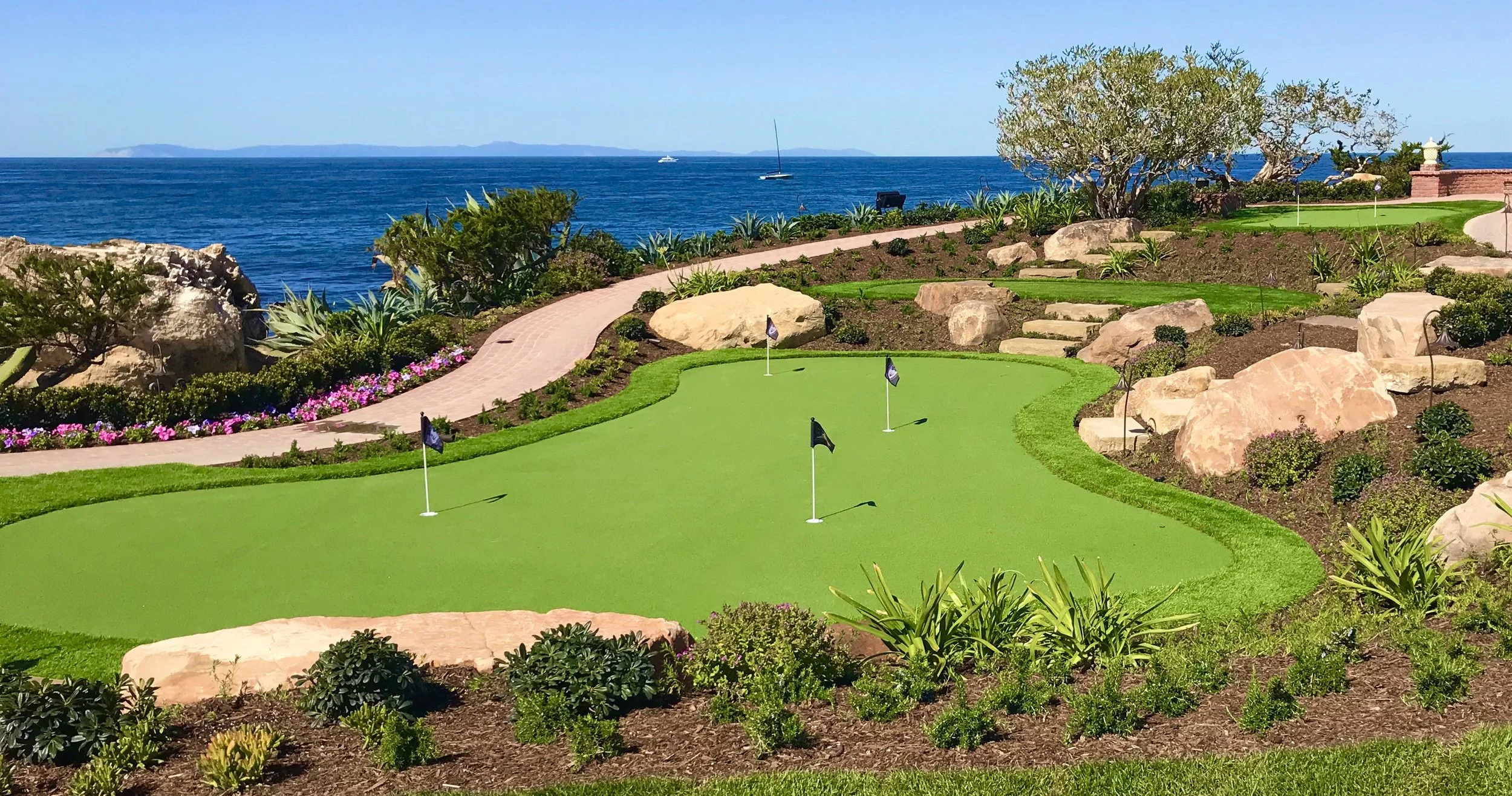 A coastal golf course with a green, flags, and rocks, overlooking the ocean with sailboats and islands in the distance.