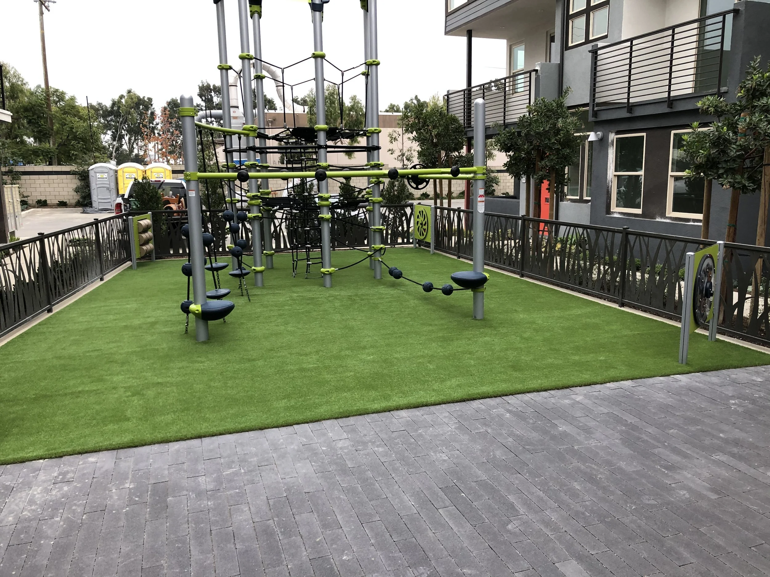 playground with green artificial turf under play equipment surrounded by a black metal fence, bushes, trees and a building on the right side.