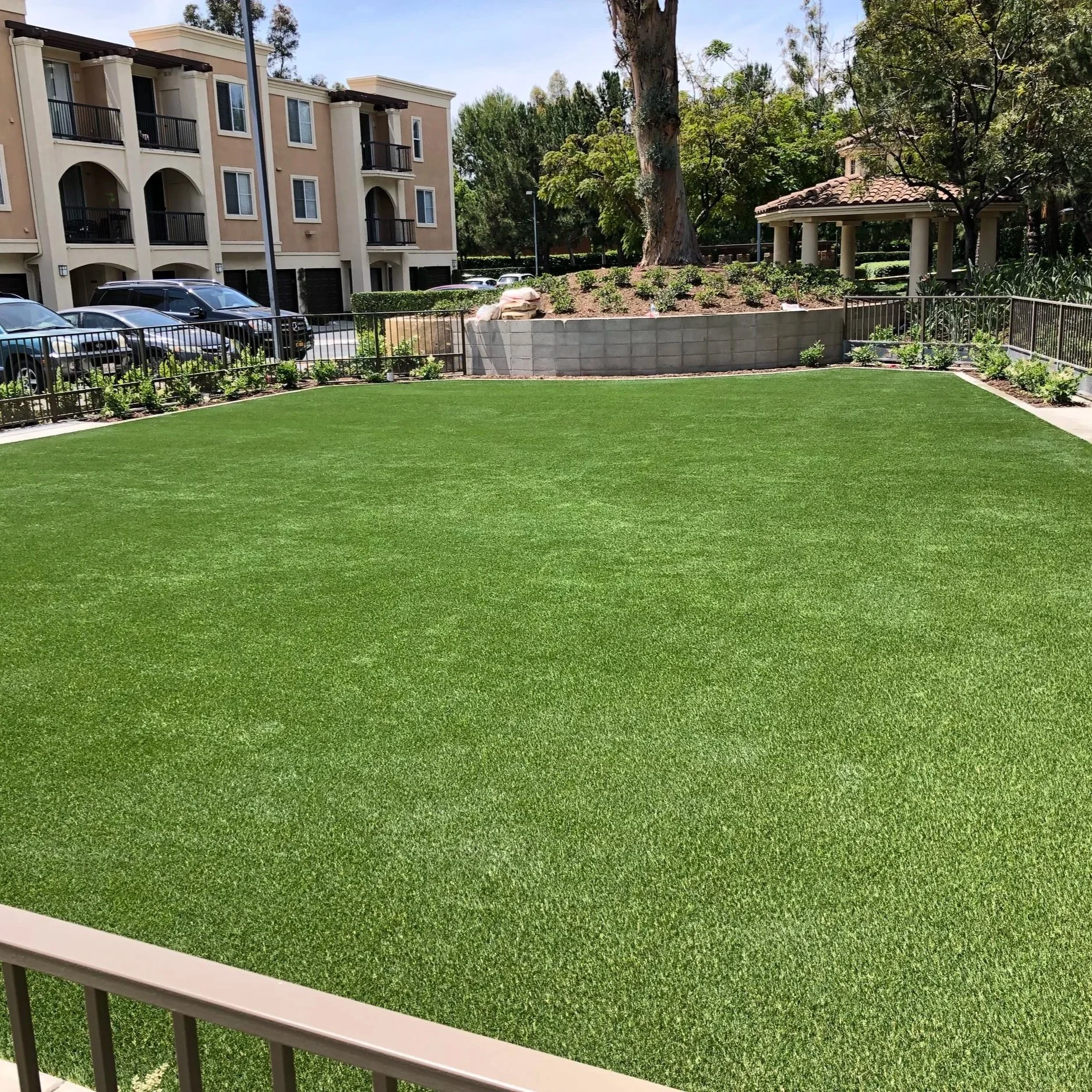 View of a furnished apartment complex with a parking lot, a lush green lawn in the foreground, trees, and a small gazebo on the right.