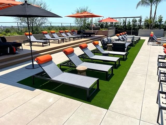 Poolside lounge chairs with orange headrests under umbrellas, placed on artificial grass and concrete, with a view of trees and a clear sky.