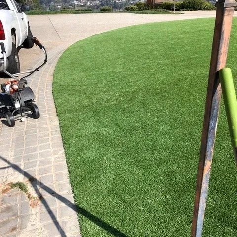 Close-up of a manicured grassy area with a curved edge, next to a paved walkway, with a mower and a vehicle in the background.