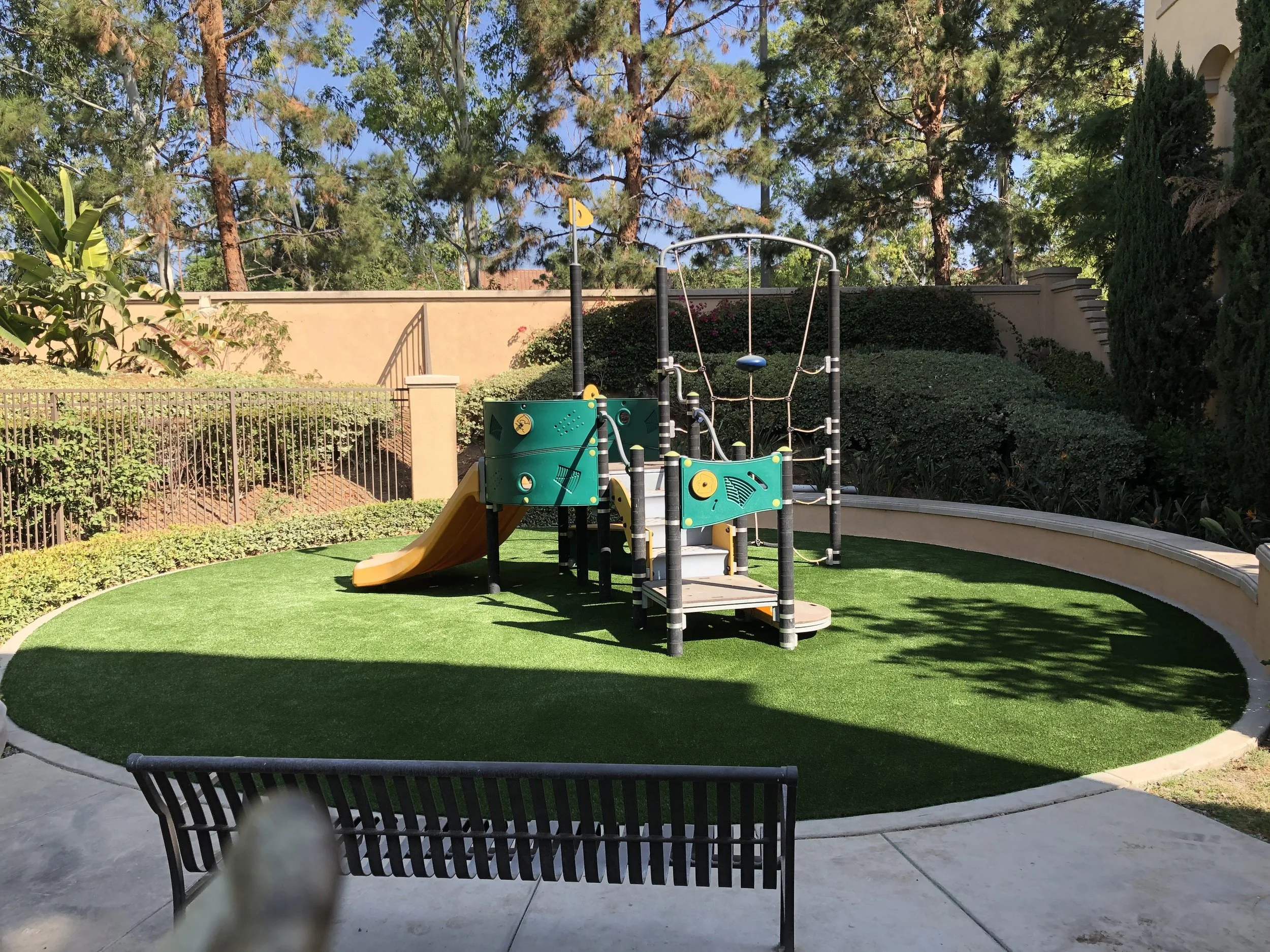 Playground with green artificial turf, climbing and sliding equipment surrounded by a low concrete wall and bushes, trees and a park bench