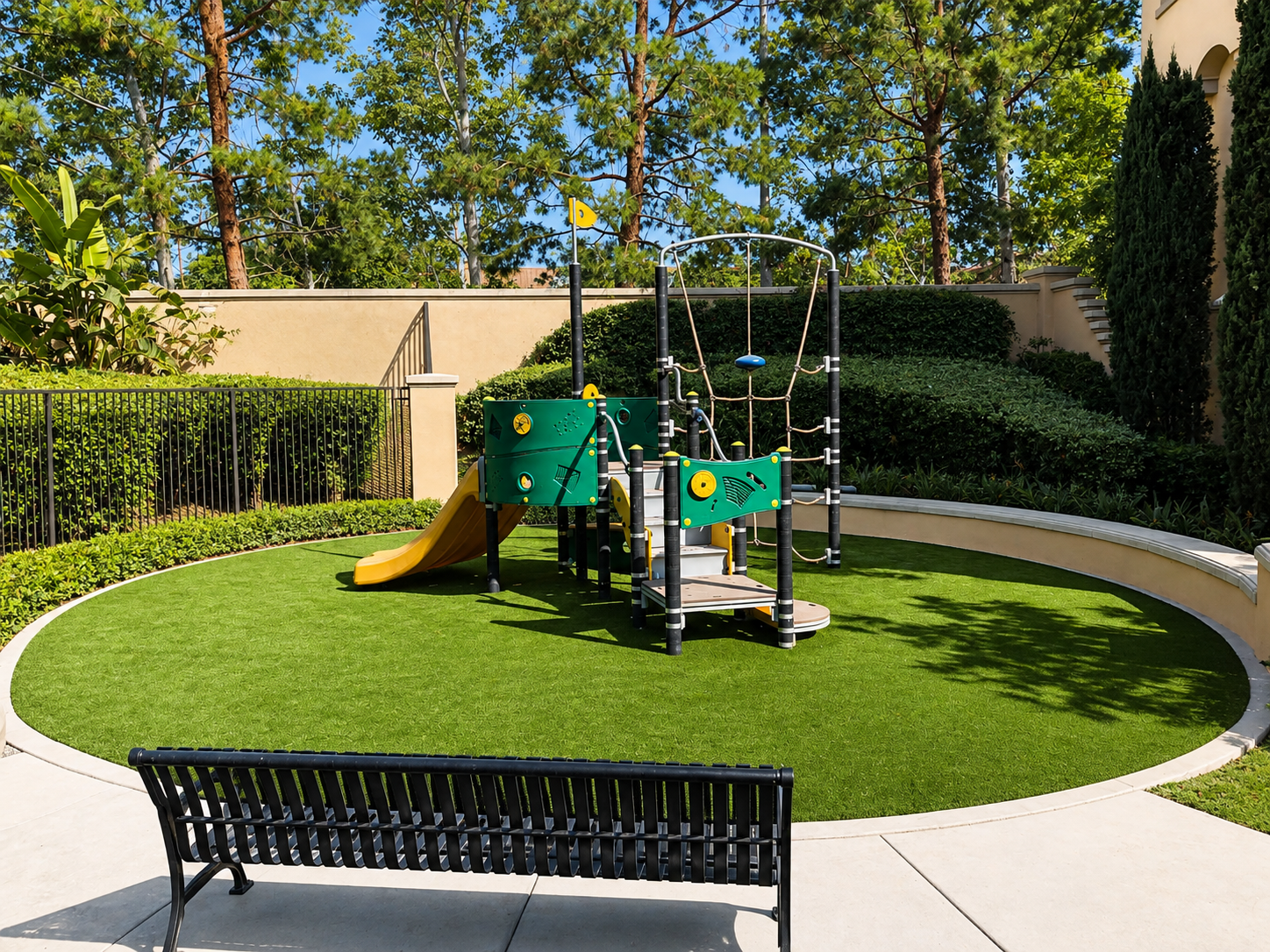 Children's playground with a small slide, climbing structure, and artificial grass, surrounded by a black fence and greenery, in a sunny outdoor setting.