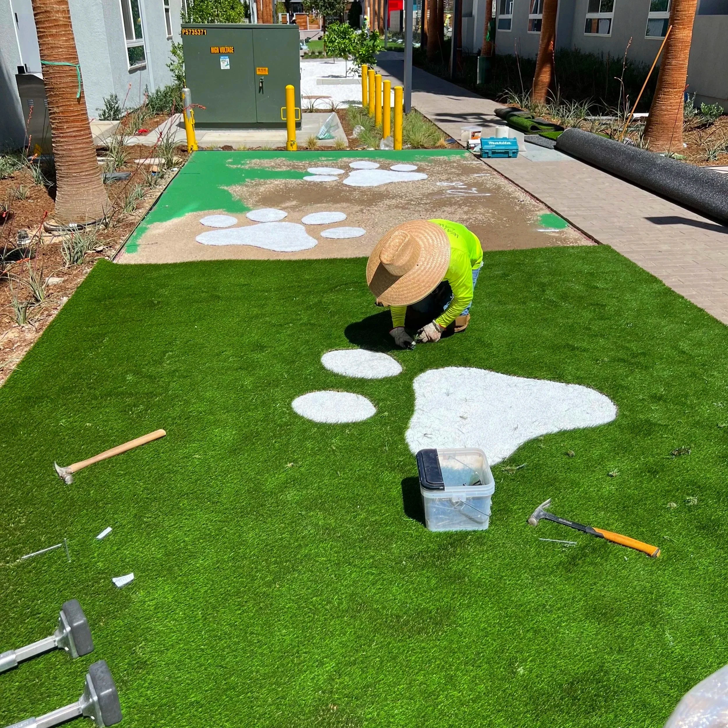 A worker installing artificial turf with white paw print designs on a landscaped area next to a sidewalk, with tools and equipment nearby.