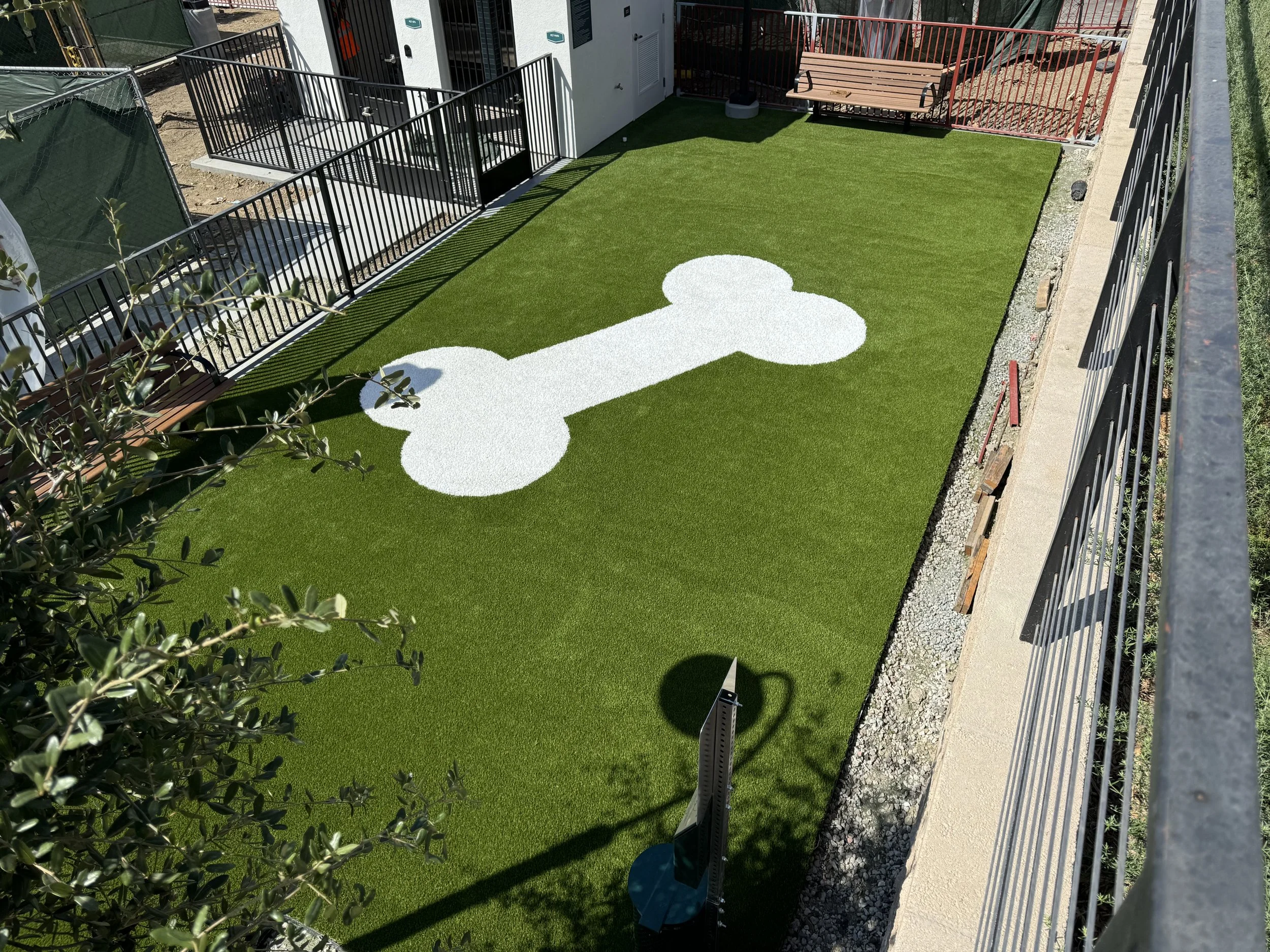 Dog park with green artificial grass featuring a white bone shape design inlayed in the green artificial turf, surrounded by black metal fencing and next to a building with utility units. There is a bench on the right and a potted plant on the left.