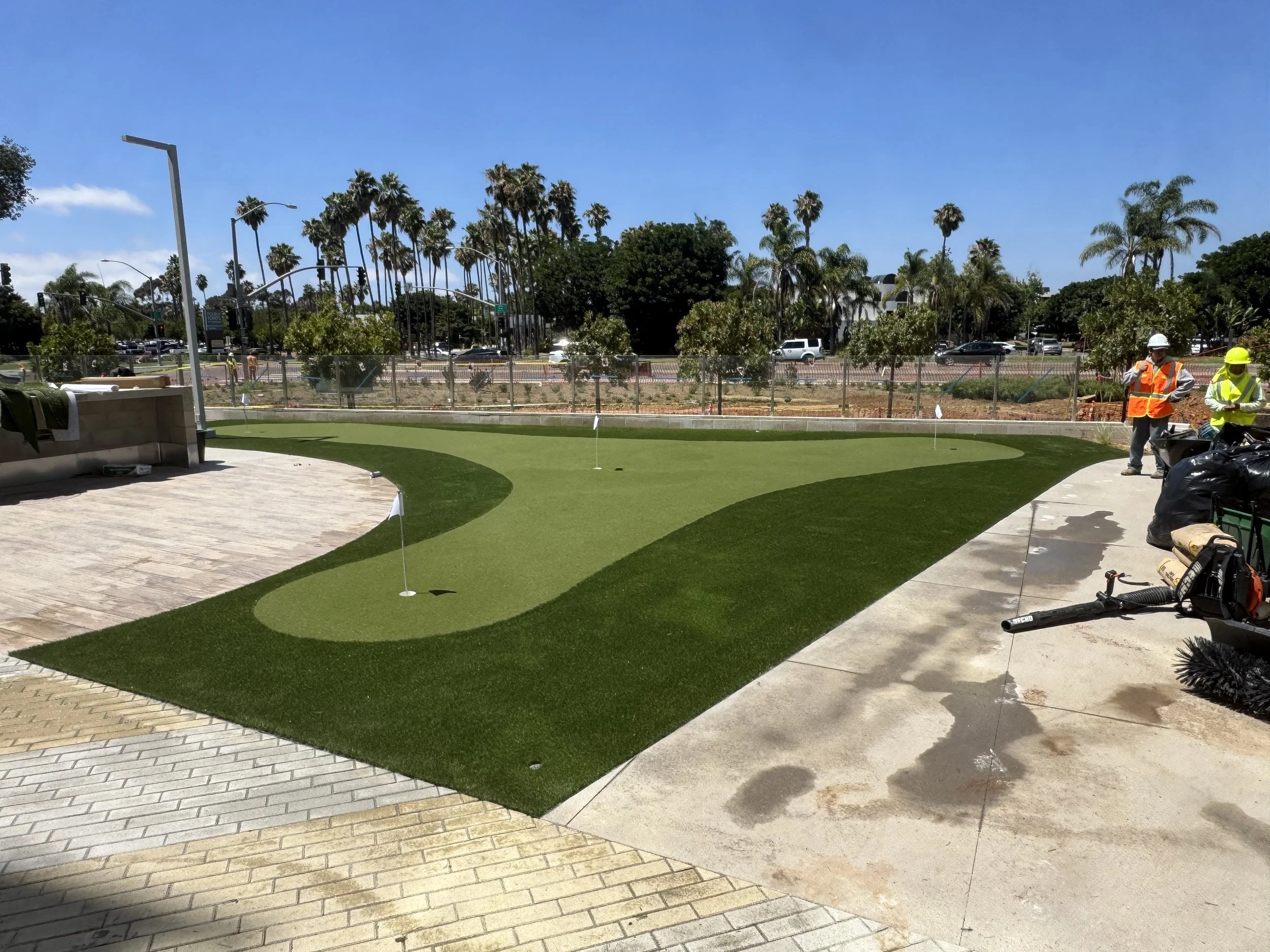 A golf course under construction with workers in safety vests and helmets, sand traps and a green in the foreground, and palm trees and a busy street in the background.