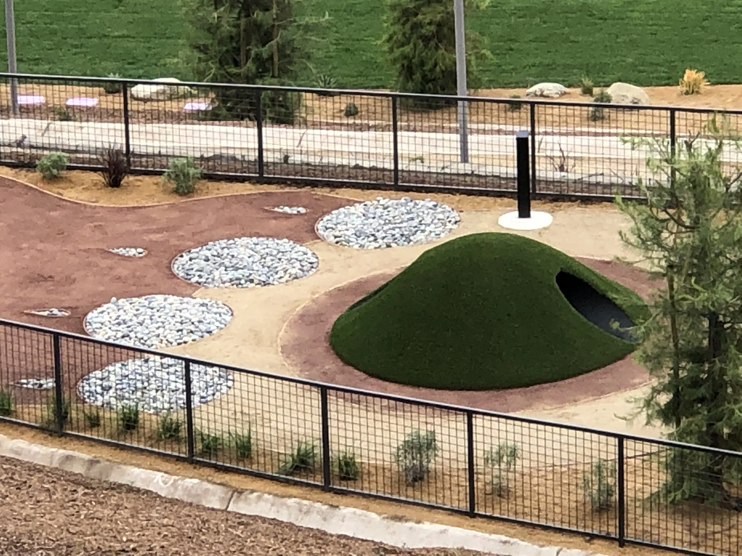 Dog park with a green artificial turf covered play mound with tunnel and paw print design made of grey river rock, enclosed with black metal fencing