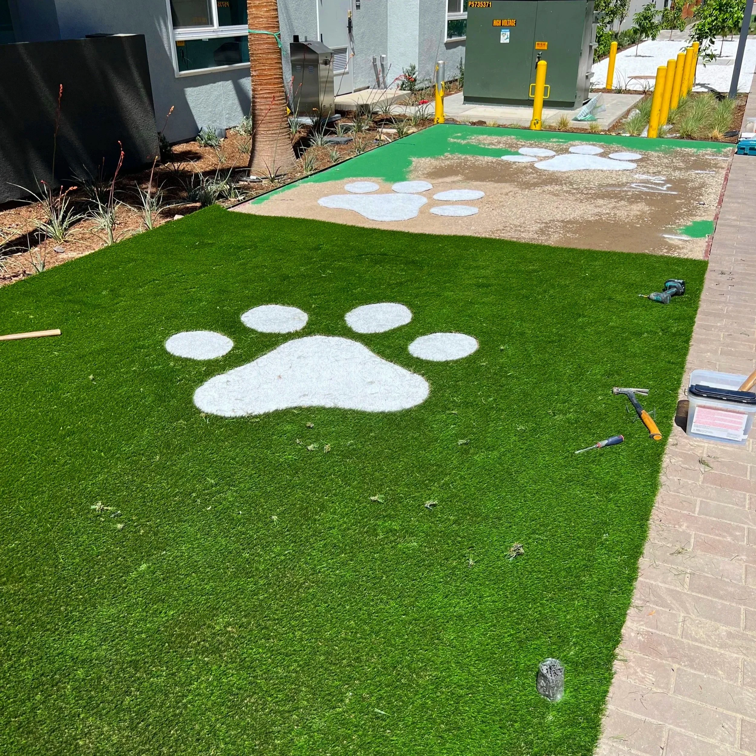 Outside area of building with green artificial turf, cement pathway, small plants, a tree, utility boxes, and yellow bollards. White paw print painted on turf and cement, ongoing landscaping or construction work.