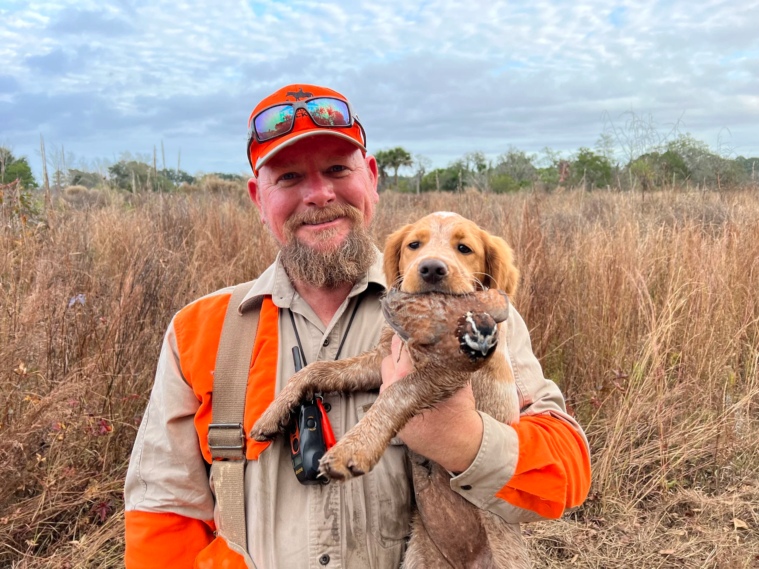 A man with a beard wearing a beige and orange outdoor hunting jacket and a red cap with sunglasses, holding a young yellow Labrador retriever puppy in a grassy field with trees and a cloudy sky in the background.