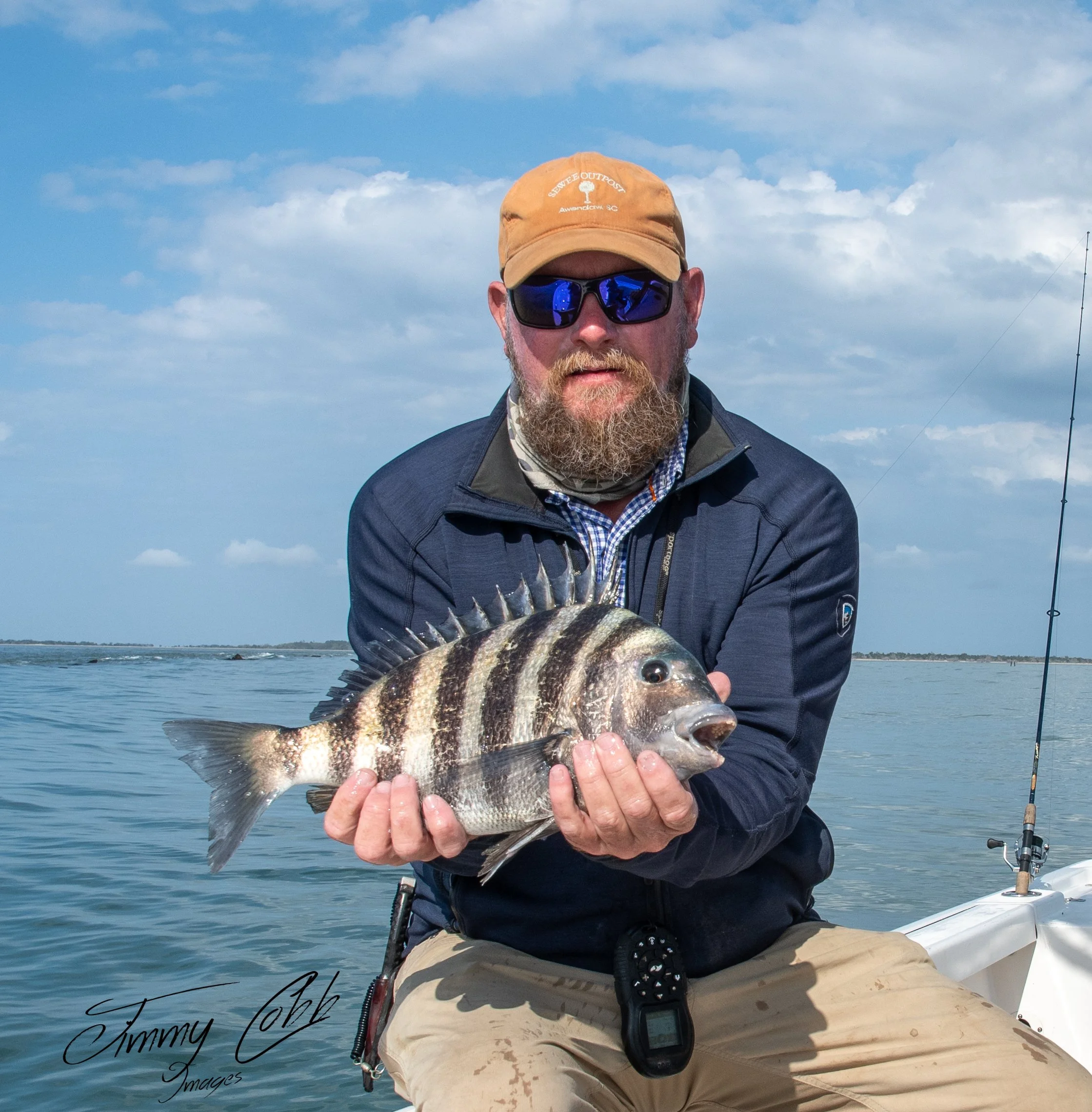 Man with a beard wearing sunglasses and a beige cap holding a striped fish while sitting on a boat in a body of water with a cloudy sky in the background.