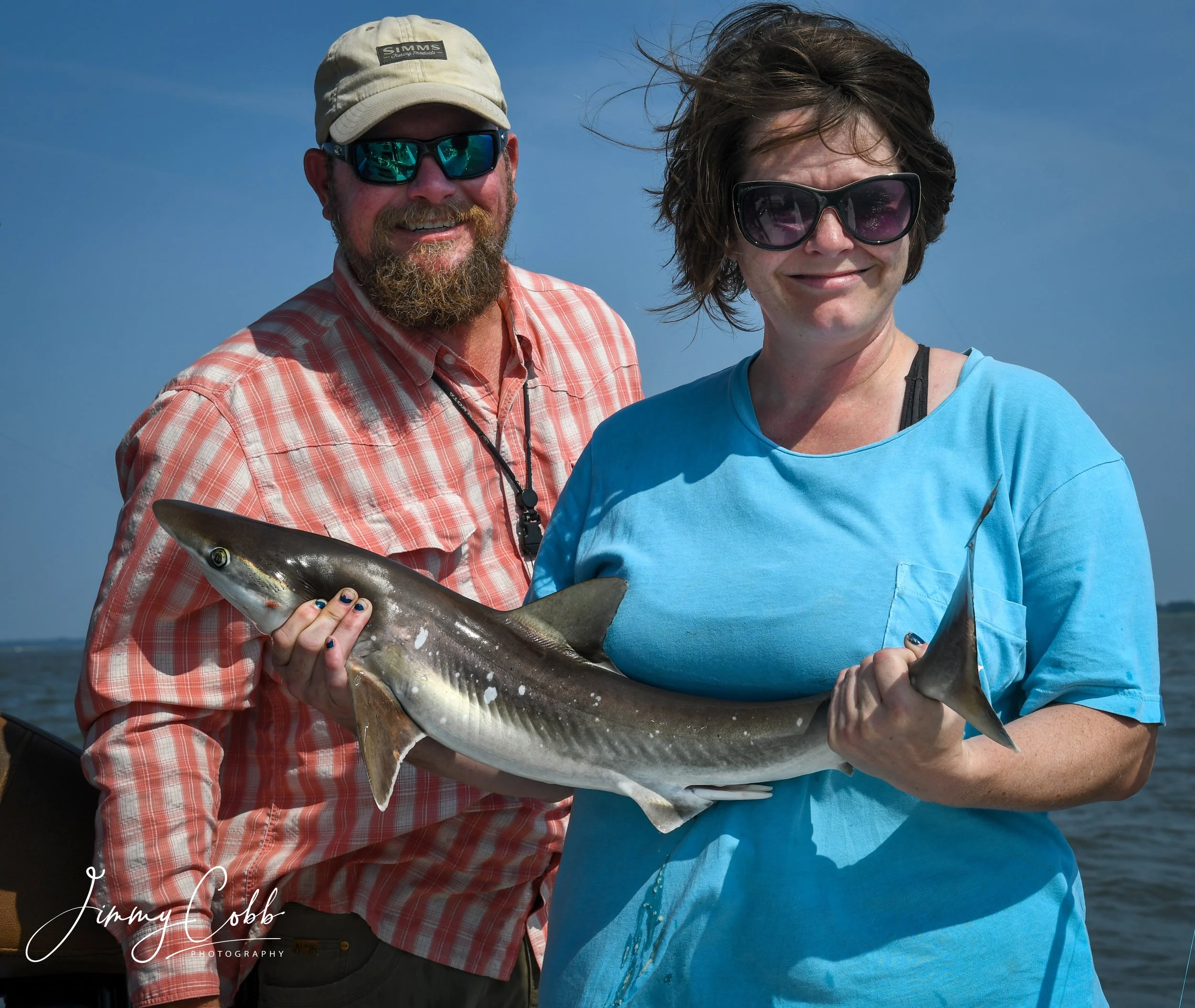 A man and a woman smiling while holding a large fish, with the man wearing sunglasses and a beige cap and the woman wearing large black sunglasses and a blue shirt, outdoors against a blue sky.
