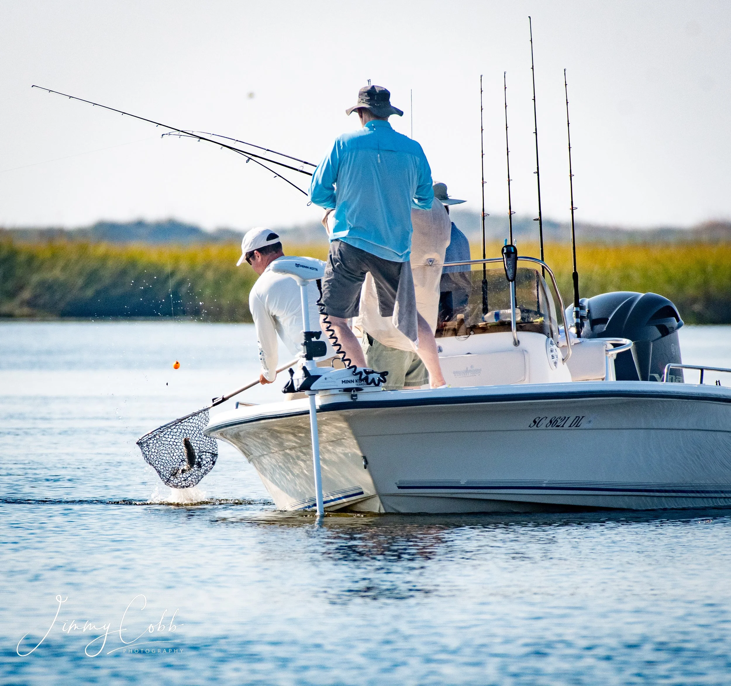 Two men in boat fishing on calm water with fishing poles, wearing hats and casual clothing.