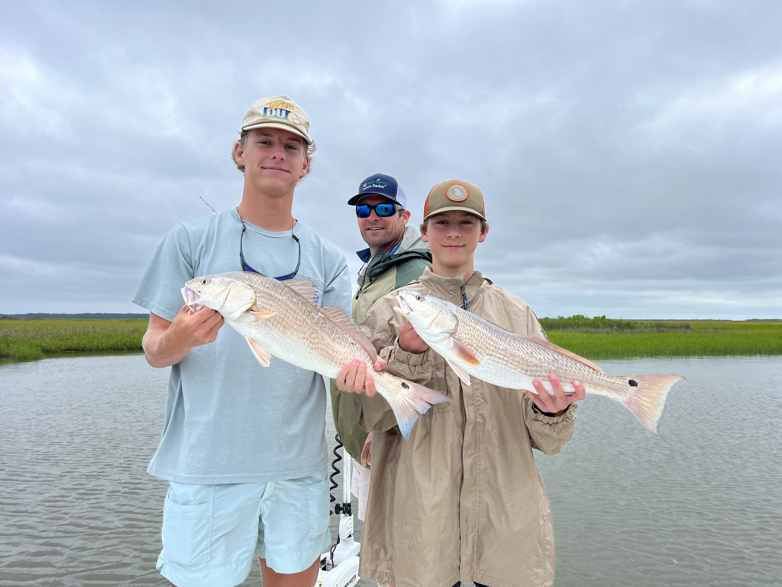 Three people standing outdoors near water, each holding a large fish they caught, smiling, under a cloudy sky.