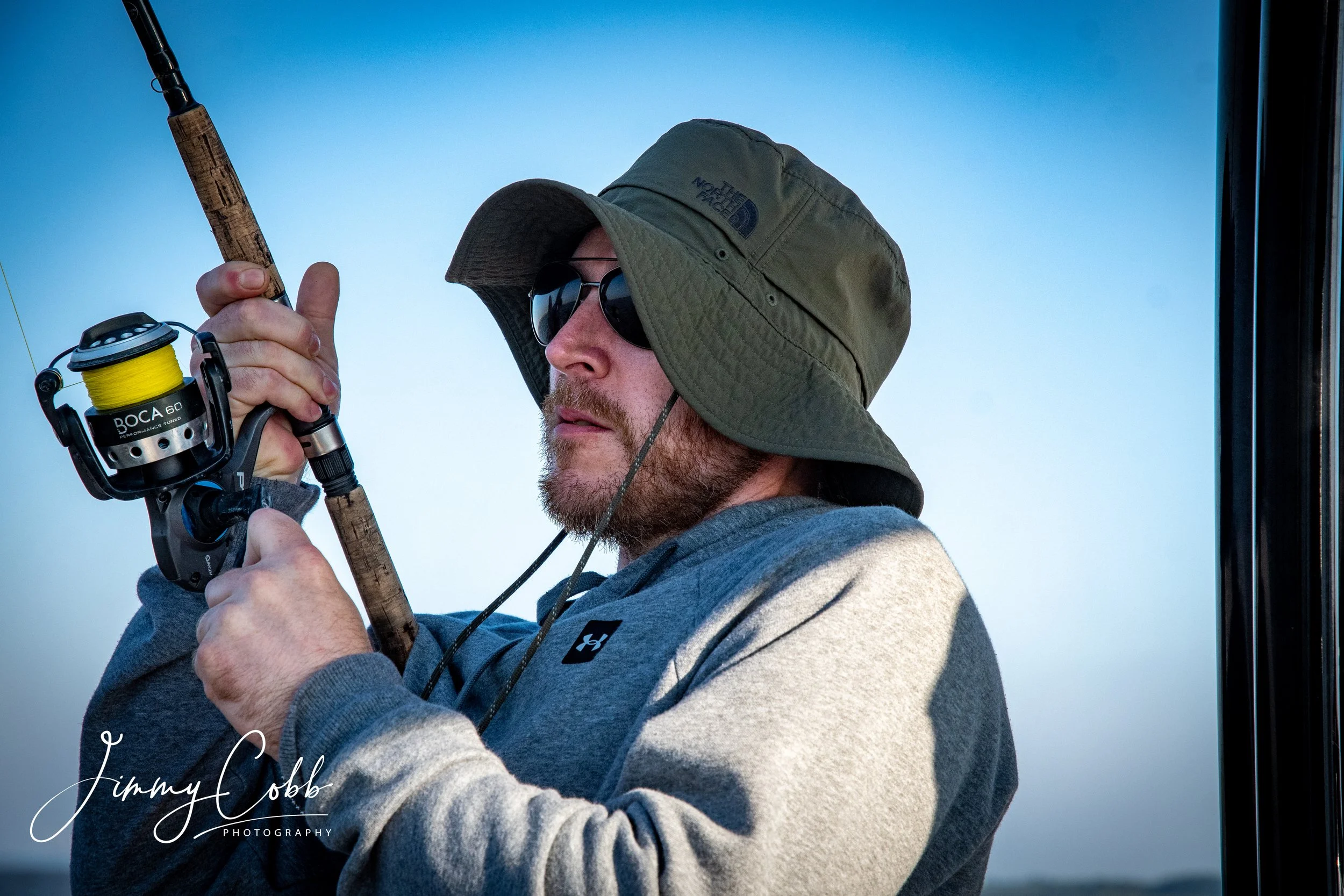 A man wearing a wide-brimmed hat, sunglasses, and a gray Under Armour hoodie holding a fishing rod with a bright yellow reel, set against a blue sky.