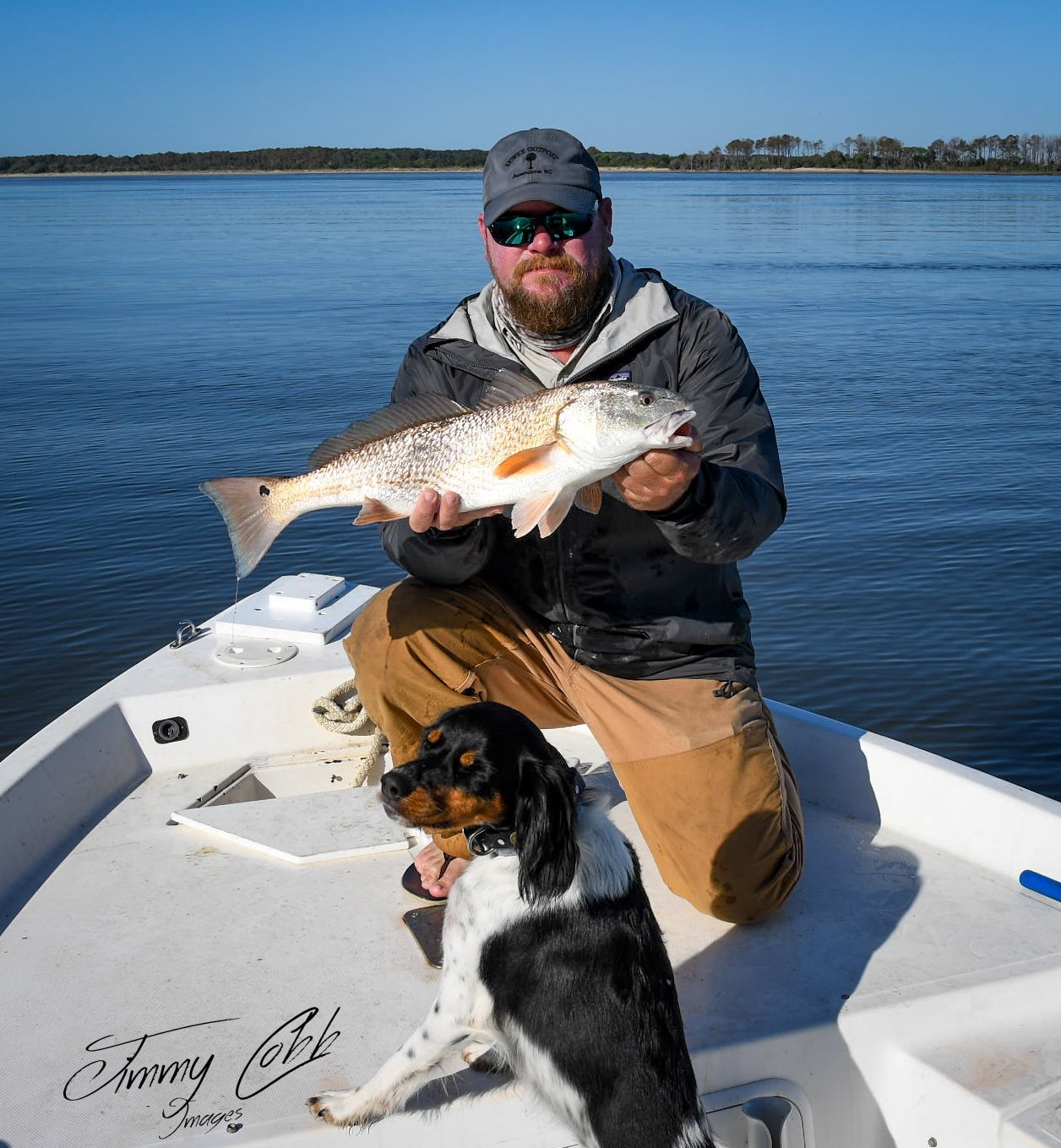 Man in sunglasses holding a large fish on a boat with a dog sitting beside him, water and distant shoreline in the background.